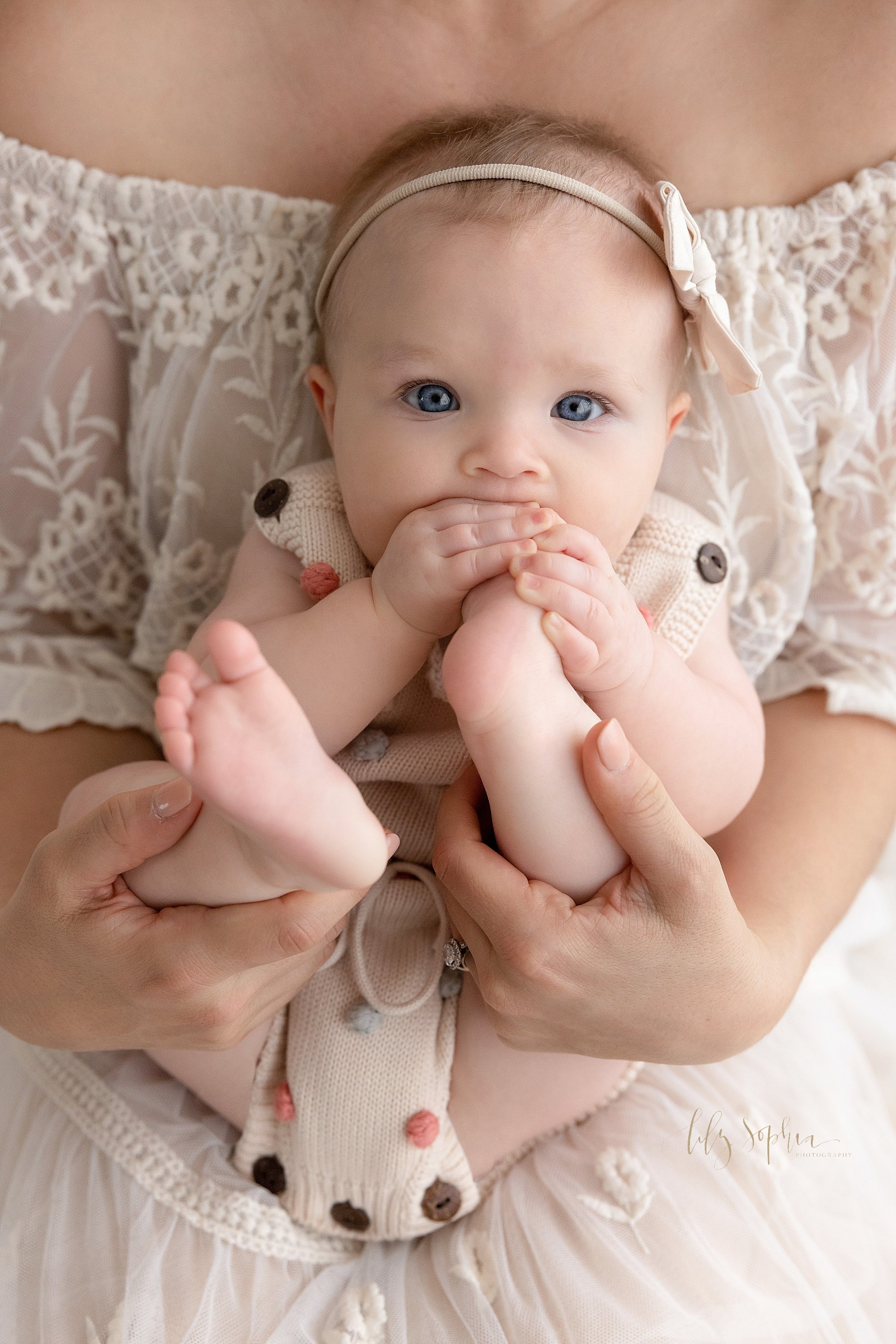  Baby milestone portrait of a baby girl sitting on her mother’s lap and grabbing her left foot with her hands to shove her foot into her mouth taken near Kirkwood in Atlanta in a photography studio that uses natural light. 