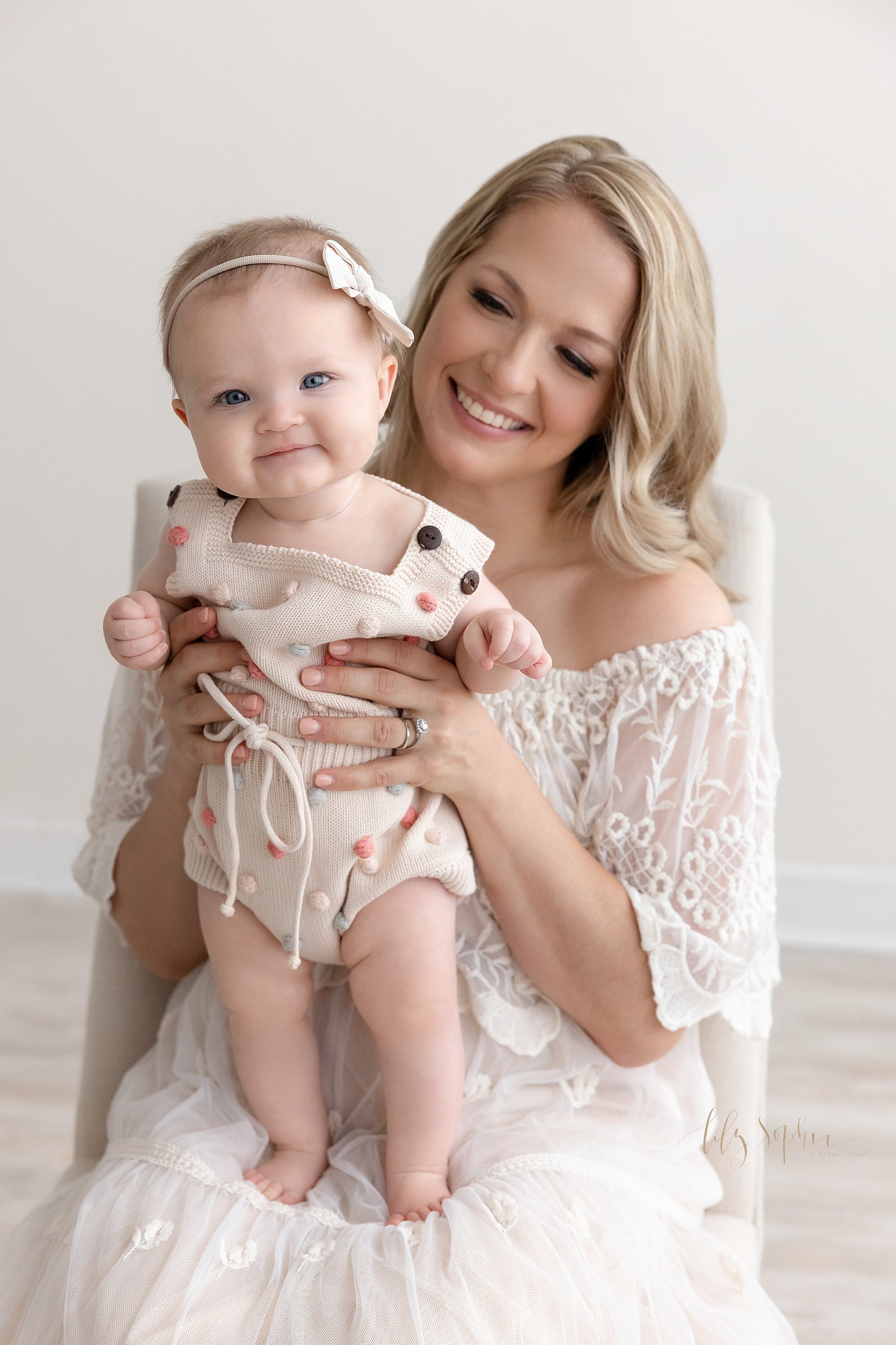  Baby photo of a baby girl proudly standing on her mother’s lap as mom looks on taken in a natural light studio near Smyrna in Atlanta, Georgia. 