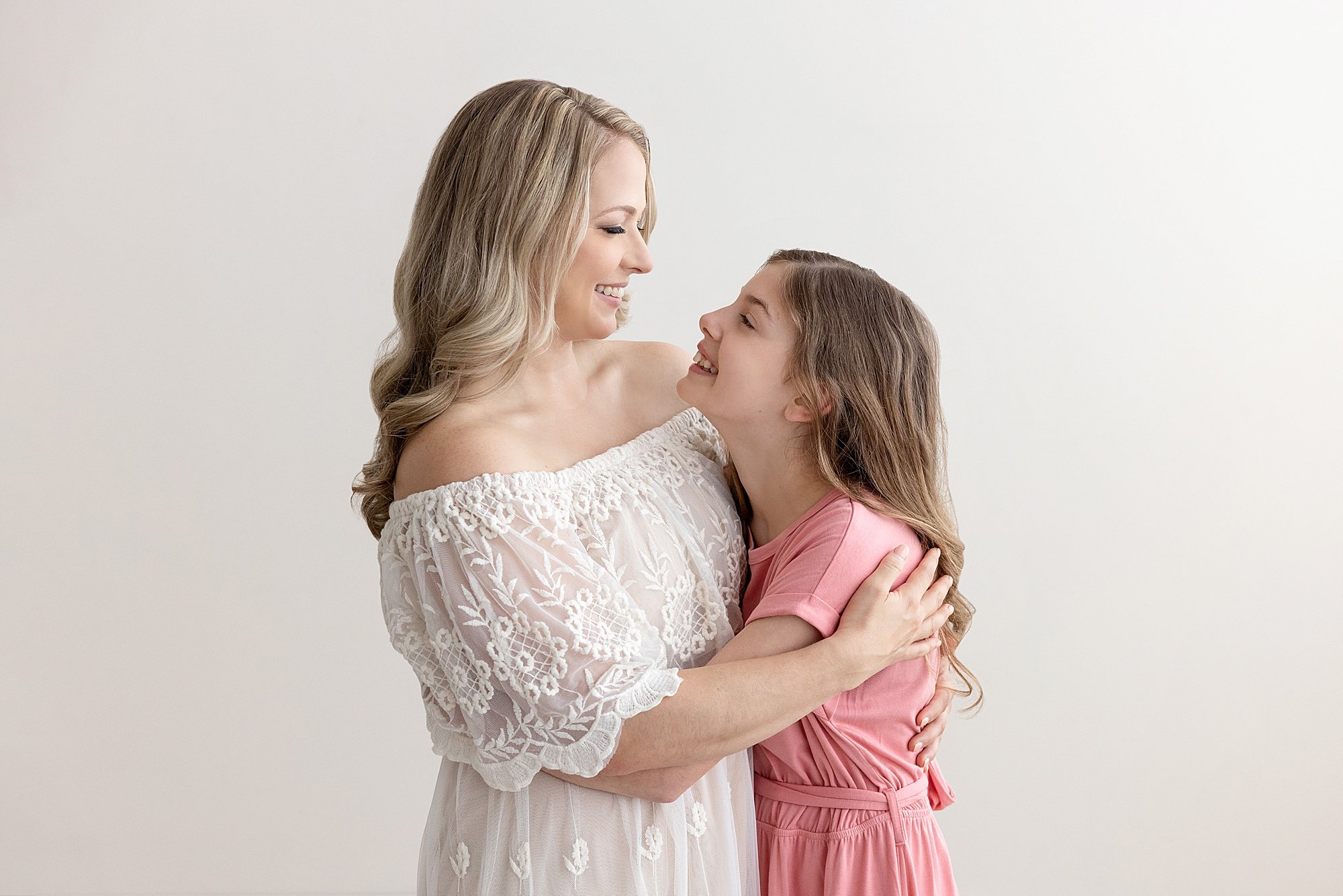  Mother and daughter family photo of a mother facing her daughter and the two of them hug one another and talk to each other as they stand next to a window streaming natural light in a studio near Decatur in Atlanta. 