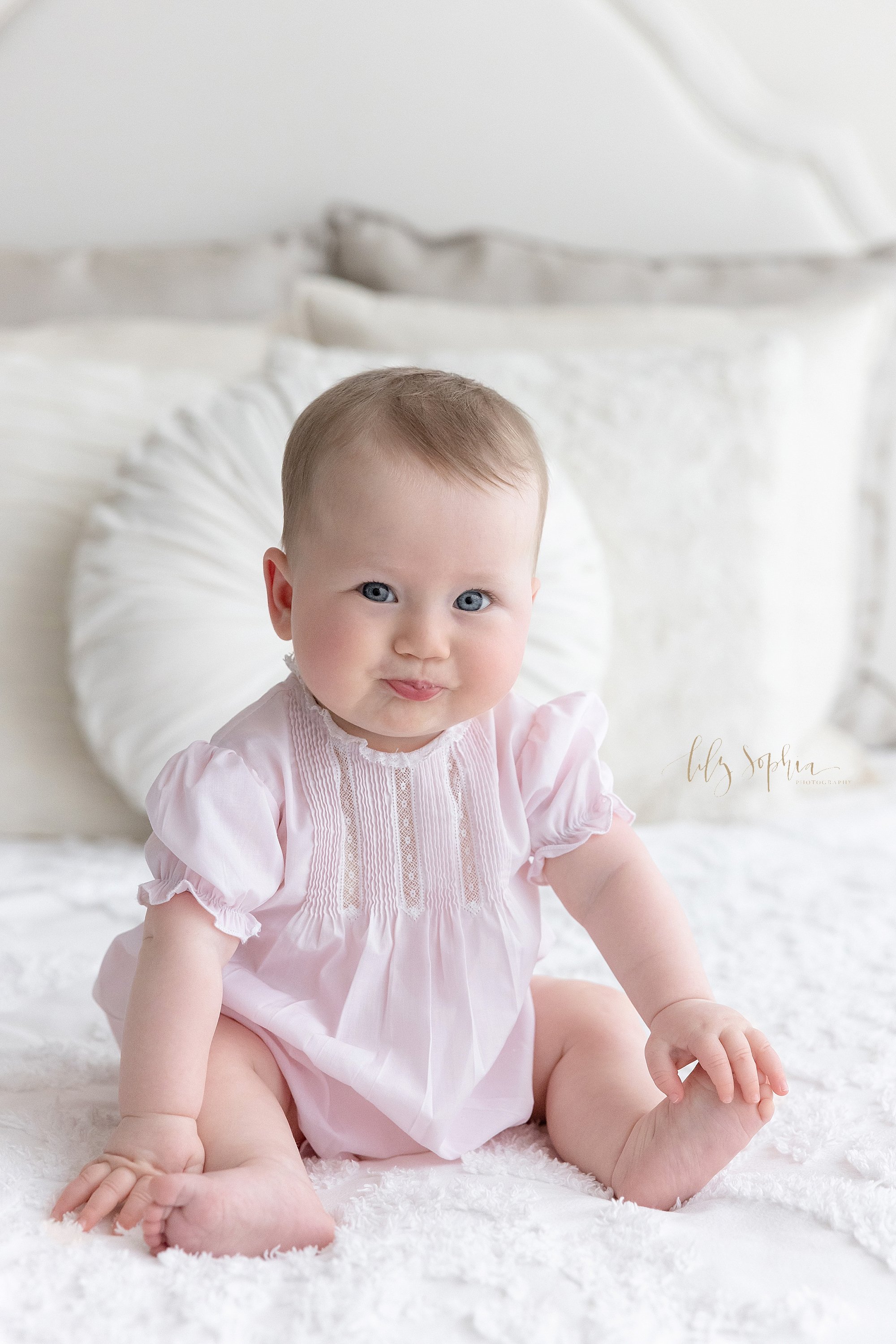  Baby photo of a six month old baby girl as she blows raspberries while sitting on a bed taken near Sandy Springs in Atlanta in a studio that uses natural light. 