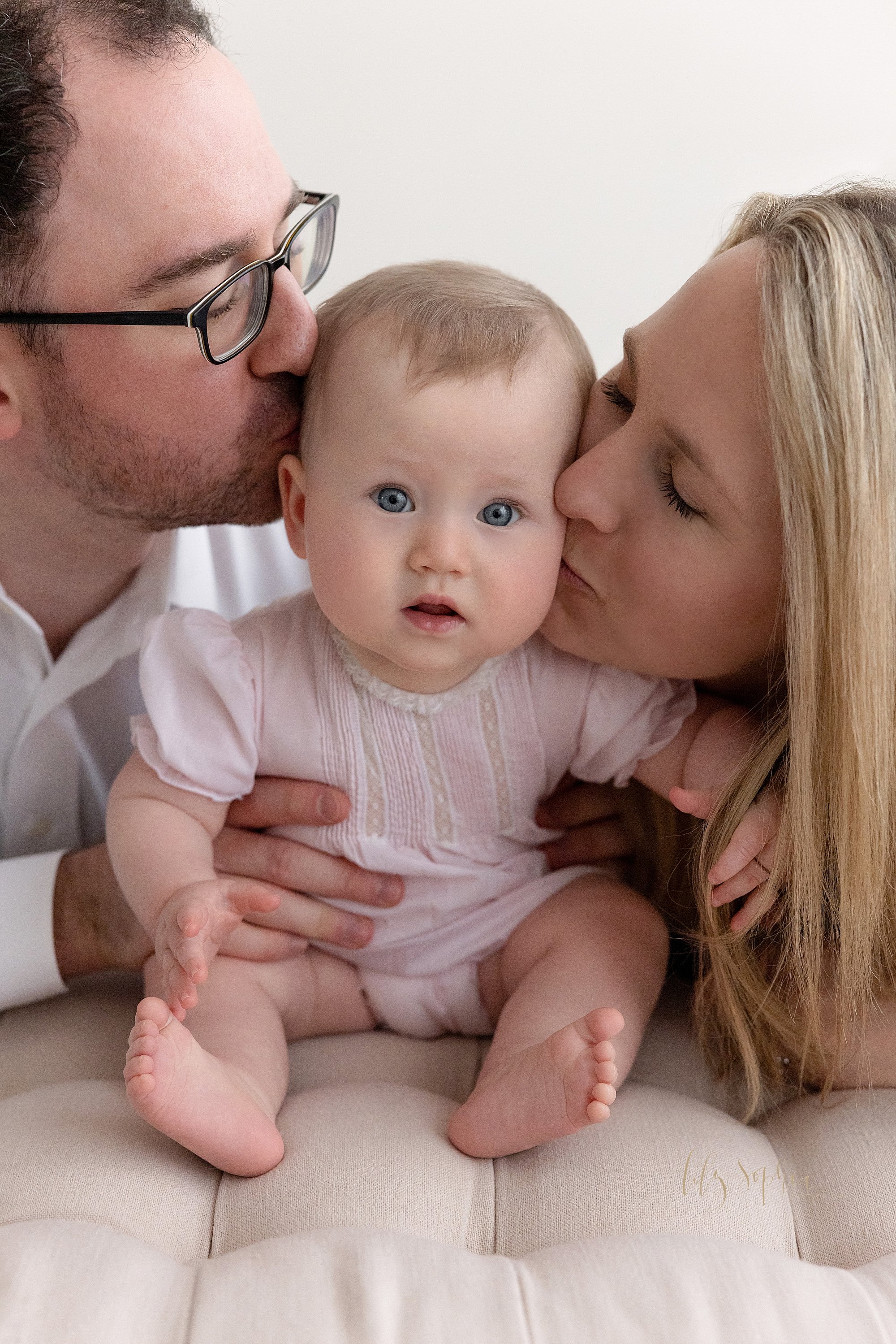  Family photo of a six month old baby girl as she sits on a tufted bench in a natural light studio with her father kneeling on her right side and mother kneeling on her left side as they kiss her on her head and her cheek respectively taken near Buck