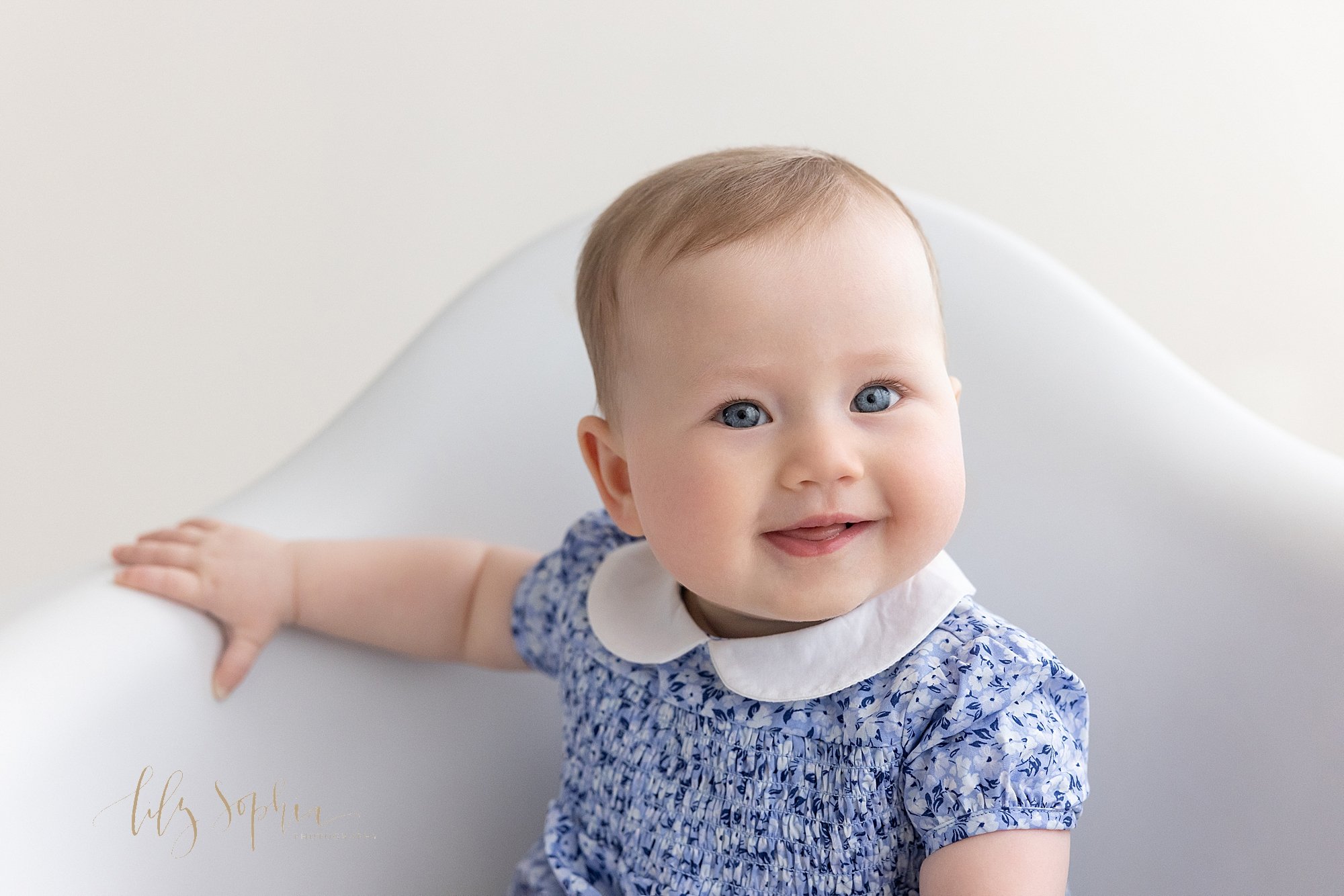  Baby milestone photo session of a six month old baby girl as she smiles while sitting in a white molded chair in a photography studio using natural light located near Oakhurst in Atlanta. 