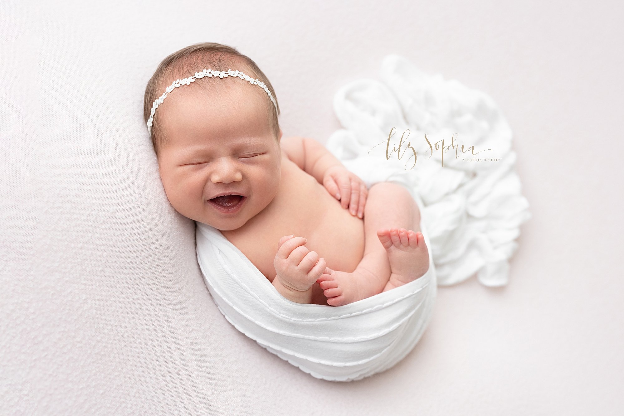  Baby photo of a laughing baby girl wearing a delicate headband in her hair as she is cradled in a blanket as she lies on her back taken near Vinings in Atlanta in a studio using natural light. 