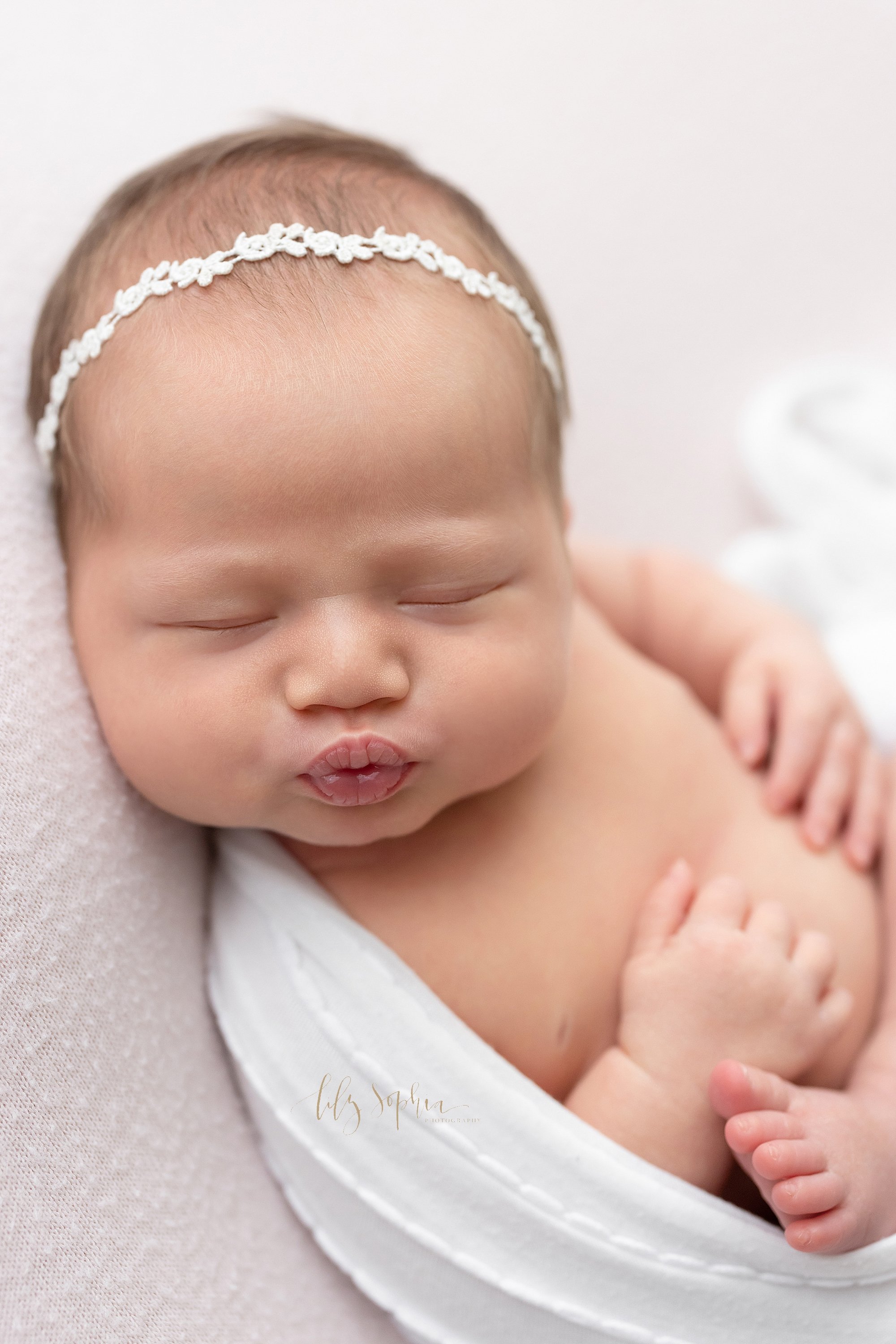  Newborn photo session of a newborn baby girl wearing a delicate headband in her wispy hair as she is cradled in a stretchy swaddle and she puckers her lips as she sleeps taken in a natural light studio near Virginia Highlands in Atlanta, Georgia. 