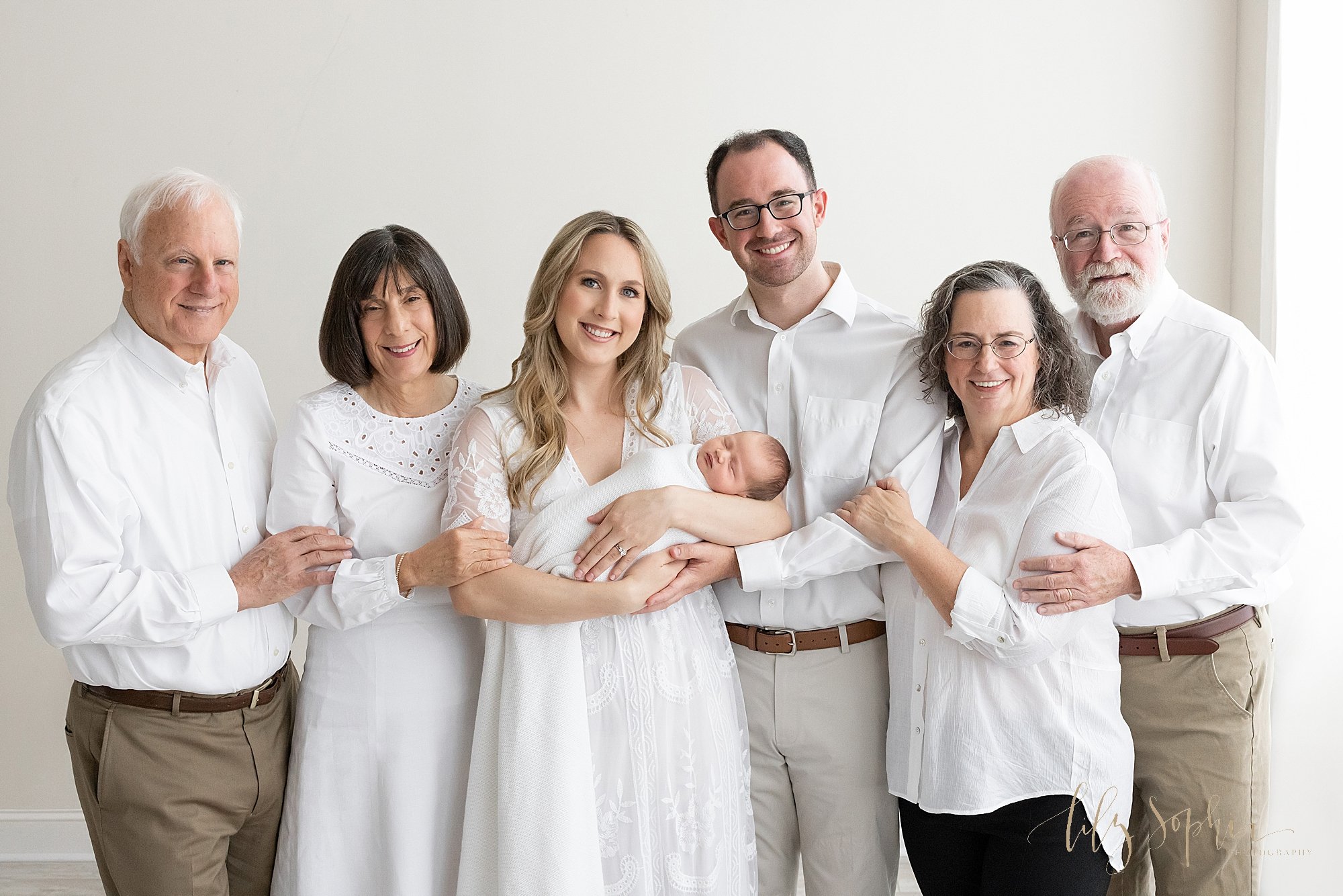  Intergenerational newborn portrait of a mother holding her newborn baby girl in her arms with her parents standing to her right side as her husband stands to her left side and his parents stand to his left side taken near Cumming in Atlanta in a stu