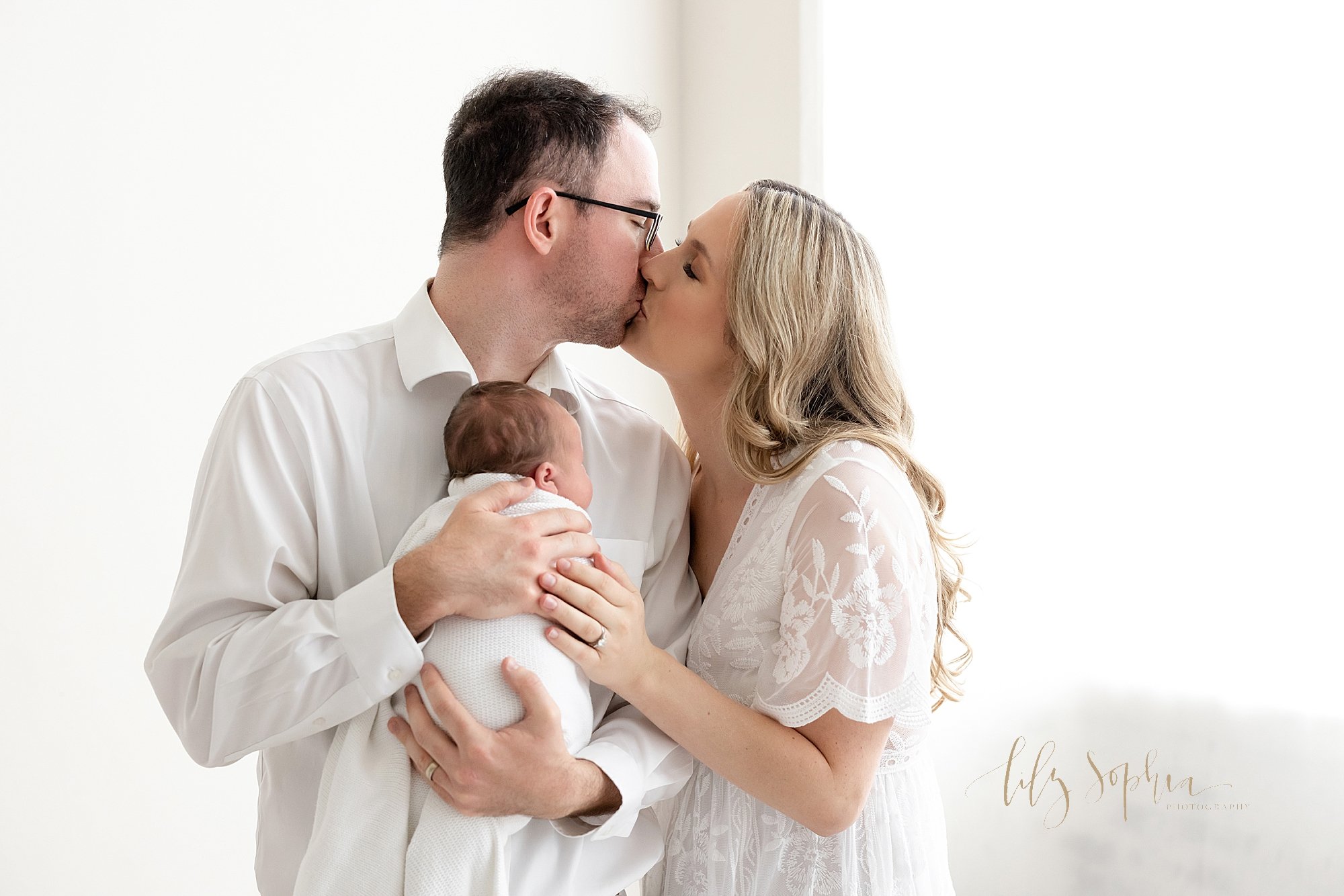  Newborn photo of a father holding his newborn baby girl against his chest as his wife stands to his right and the two kiss in front of a window streaming natural light in a photography studio near Sandy Springs in Atlanta, Georgia. 