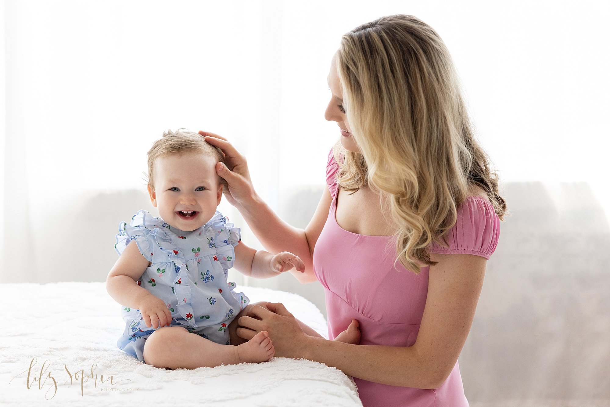  First birthday photo session of a happy one year old baby girl as she sits on a bed in front of a window streaming natural light as her mother touches her head taken in Ponce City Market in Atlanta at the Lily Sophia Photography Studio. 