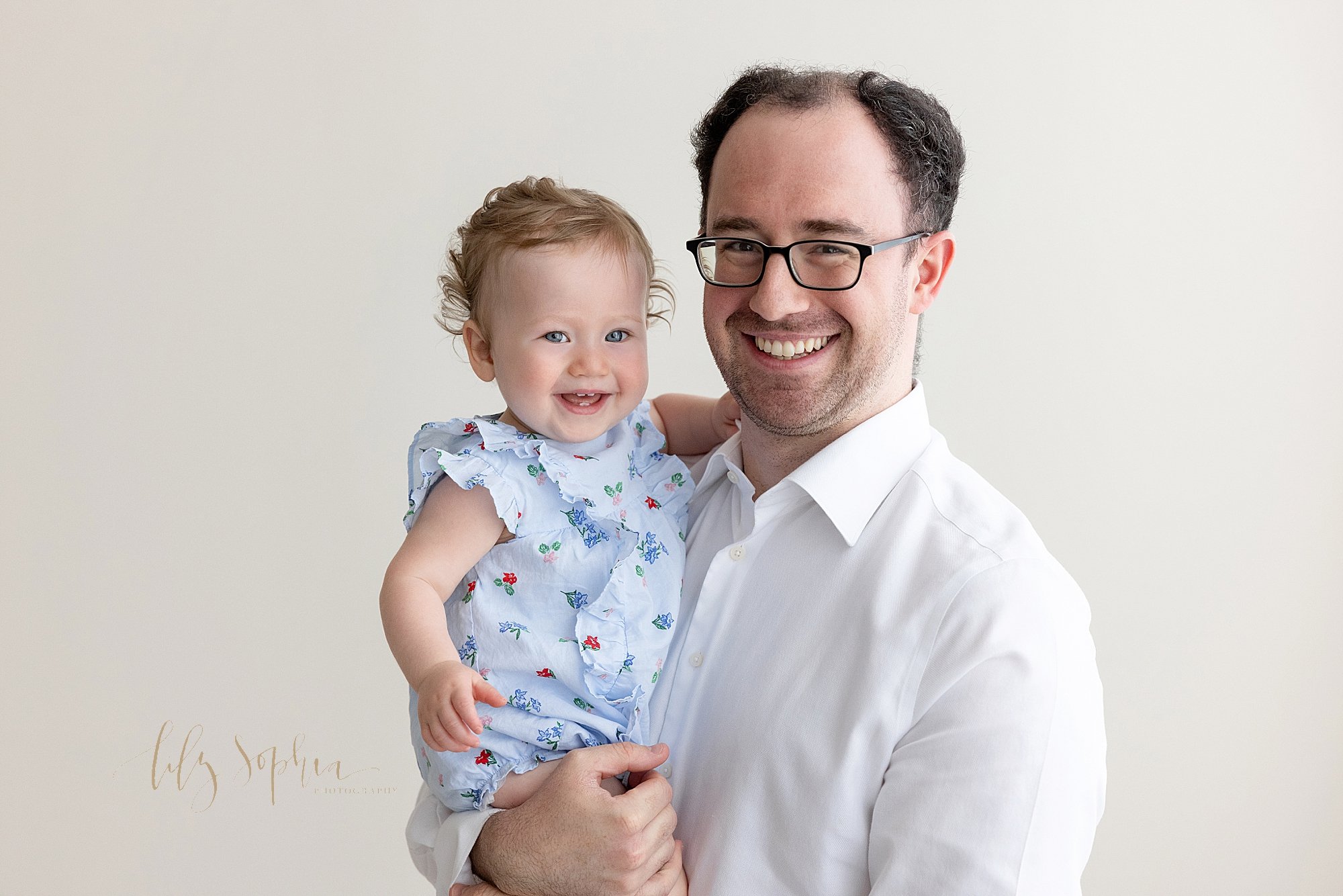  Family first birthday photo of a father holding his one year old smiling baby girl in his right arm as he stands in a natural light studio near Virginia Highlands in Atlanta.   