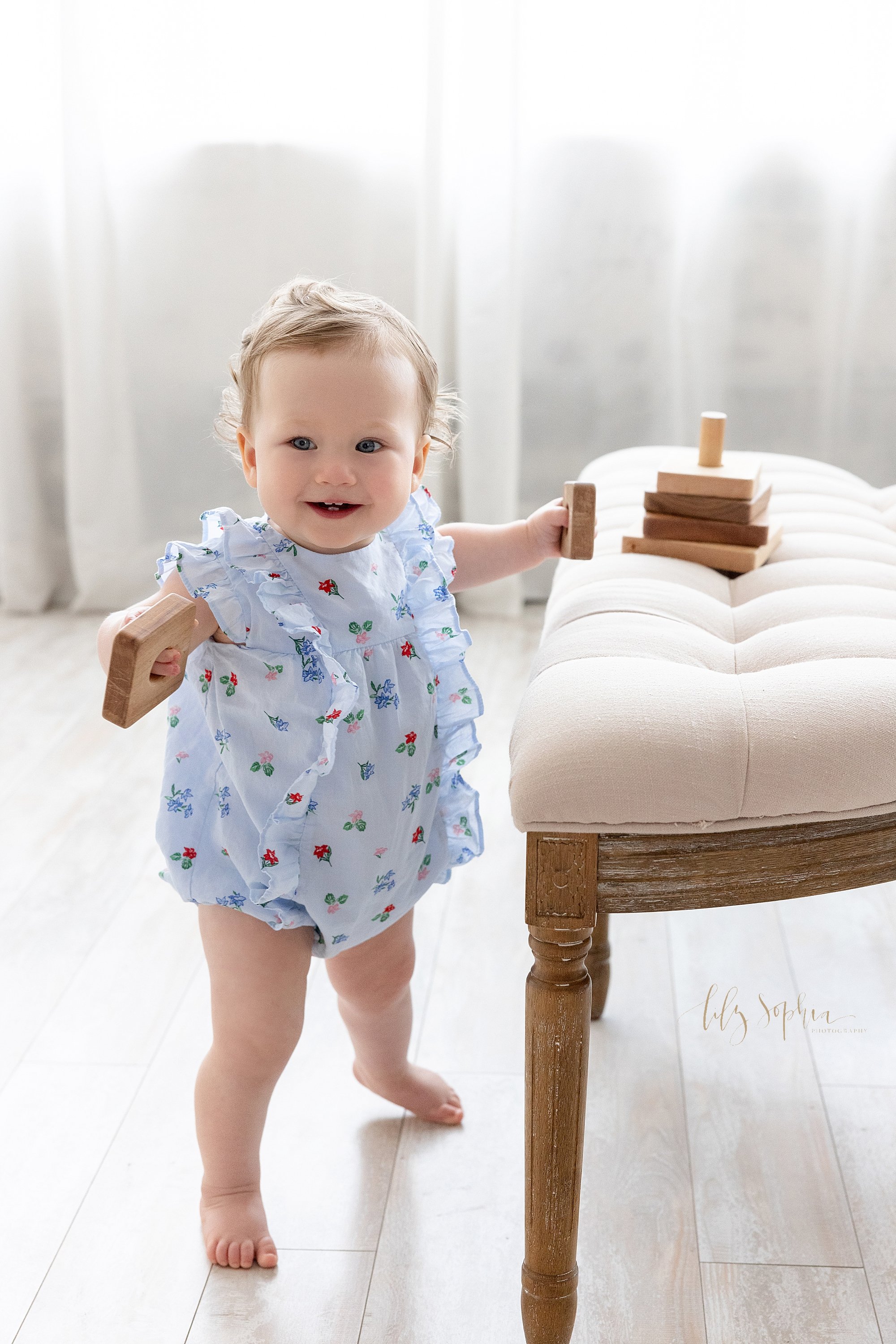  First birthday photo session of a one year old baby girl as she stands next to a tufted bench in front of a window streaming natural light as she plays with a wooden stacking toy taken near Virginia Highlands in Atlanta. 