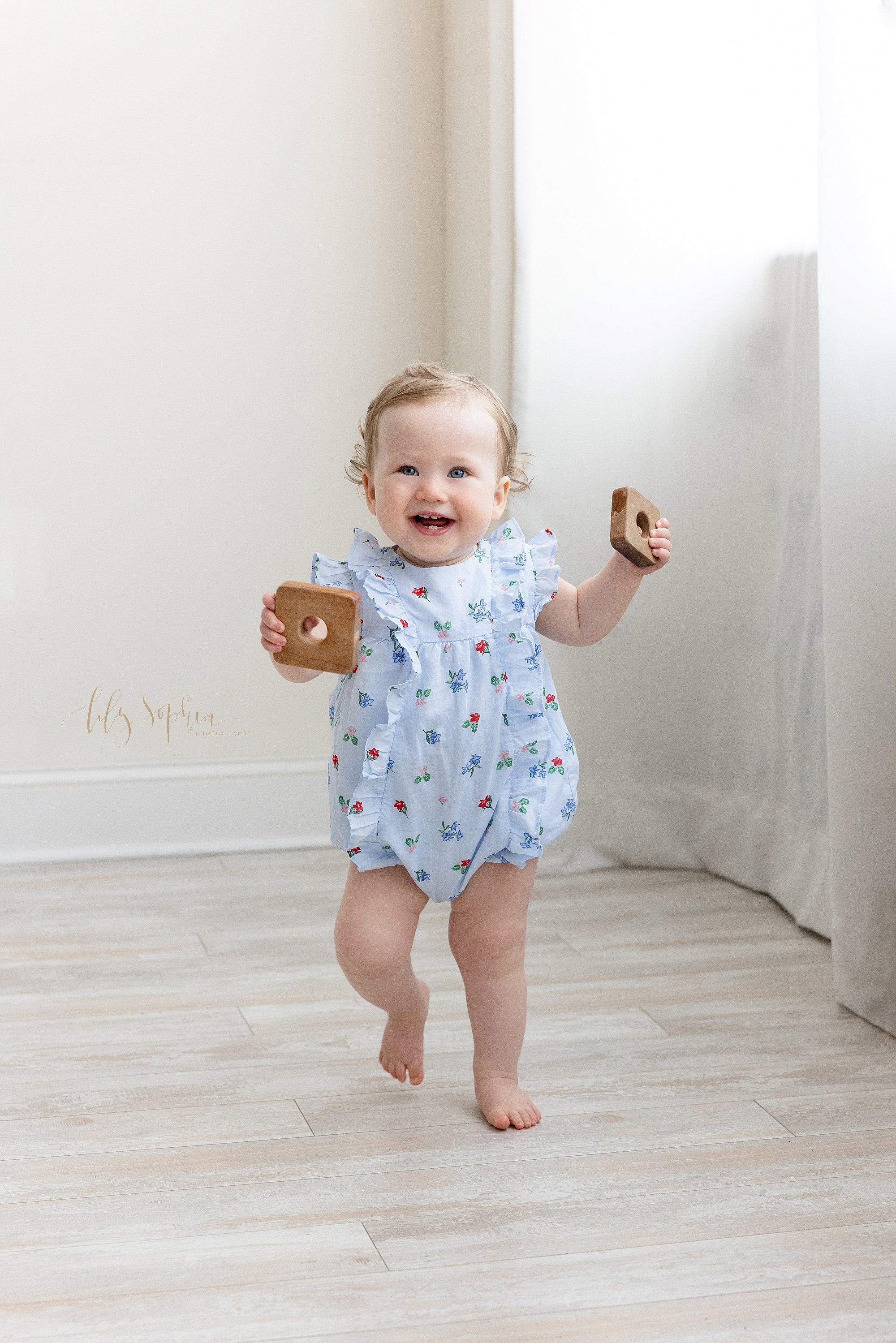  Baby photo of a smiling one year old baby girl as she walks on her own carrying wooden stacking pieces in a studio using natural light for her first birthday photo shoot taken near Poncey Highlands in Atlanta. 