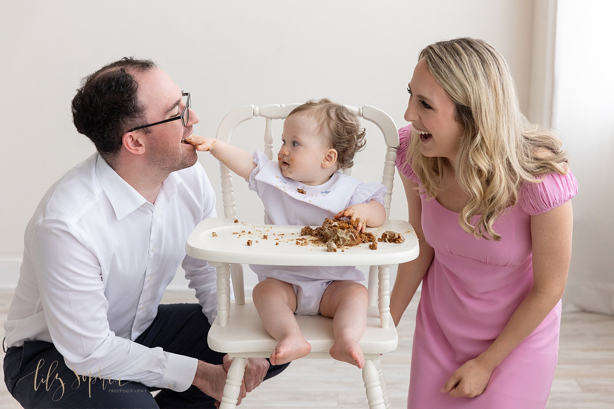  Family first birthday photo of a baby girl sitting in an antique high chair as she feeds part of her smash cake with her dad who squats on her right side and mom squats and looks on on her left side taken near Cumming in Atlanta, Georgia. 