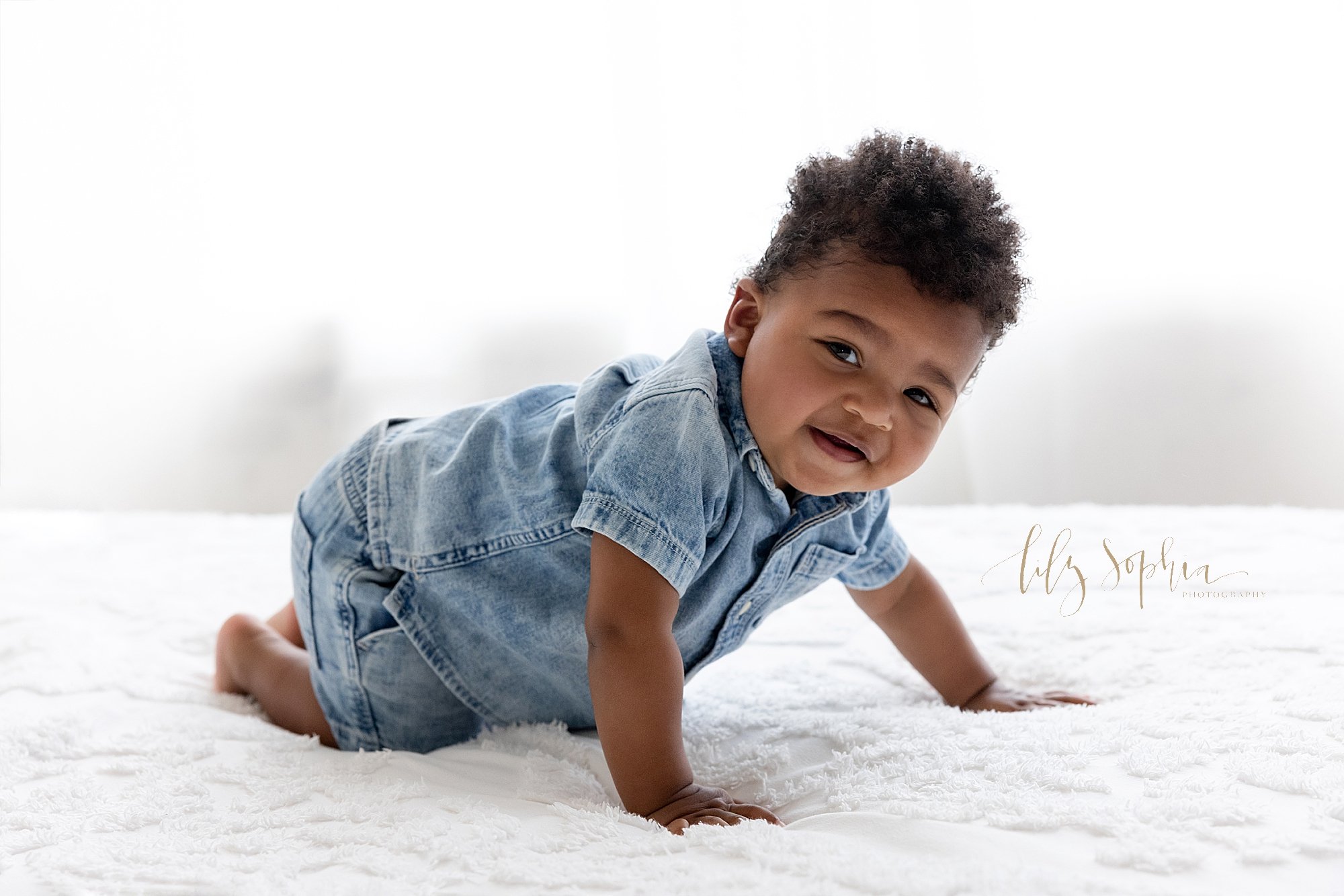  Baby portrait of an African-American baby boy as he crawls on the top of a bed with a window streaming natural light behind him taken near Old Fourth Ward in a photography studio. 