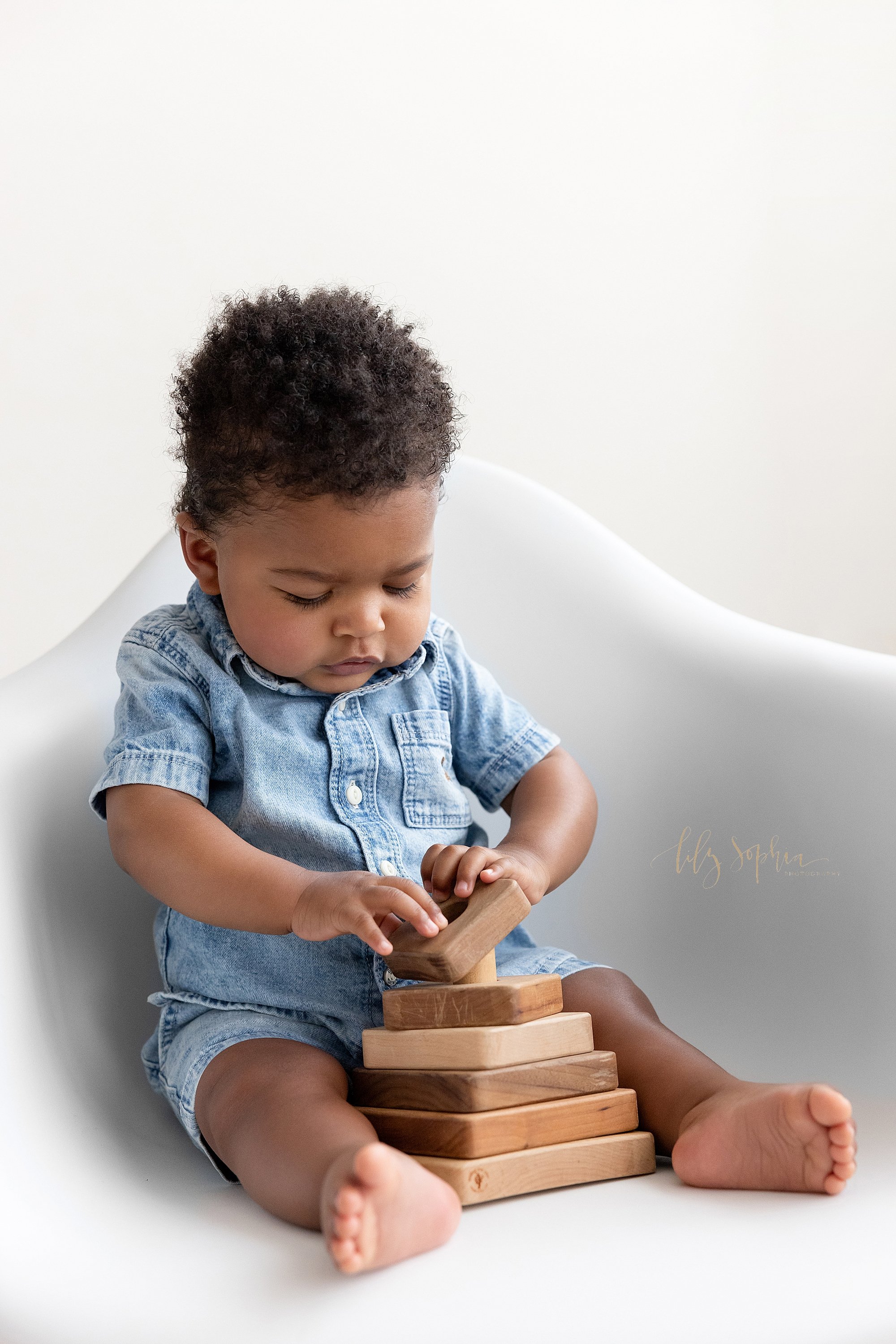  A determined African-American baby boy gets his picture taken as he sits in a white molded chair and plays with a wooden stacking toy taken in a photography studio near Smyrna in Atlanta that uses natural light. 