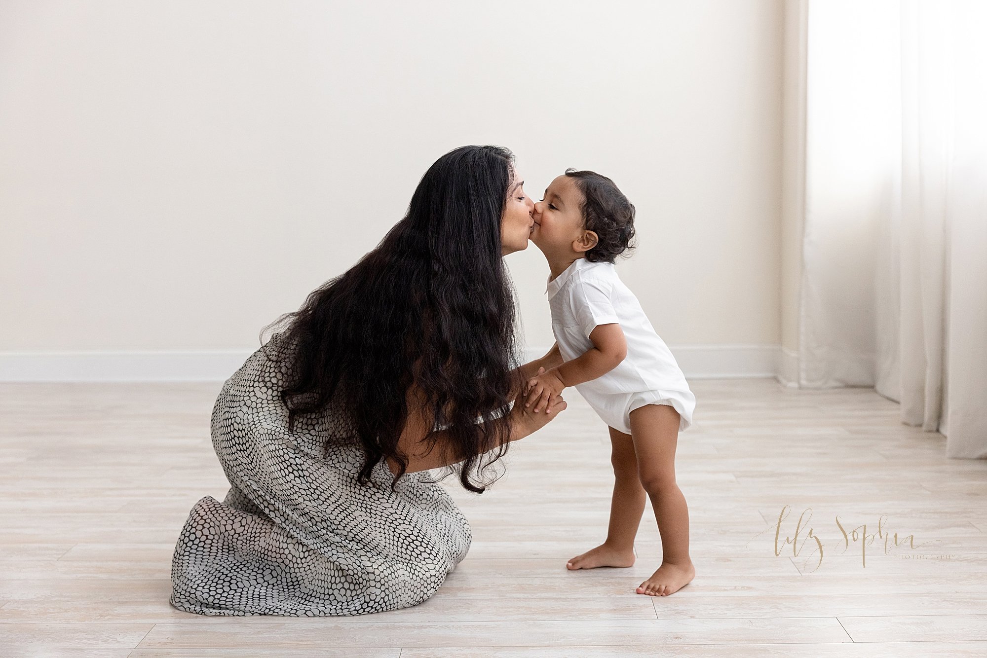  Family photo of a mother kneeling on the floor of a natural light studio with her toddler son facing her and giving his mother a kiss taken near Roswell in Atlanta. 
