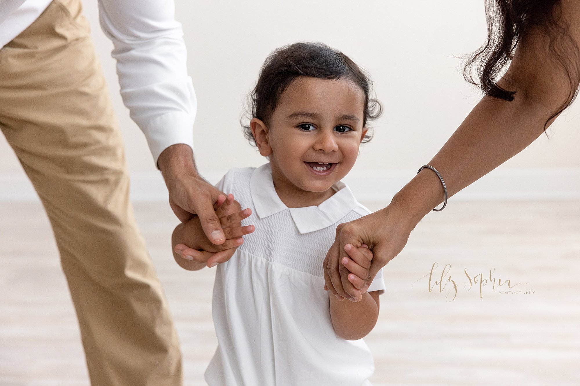  Family picture of a smiling Indian boy showing his tiny teeth as he runs holding his mom and dad’s hands in a photography studio near Virginia Highlands in Atlanta that uses natural light. 