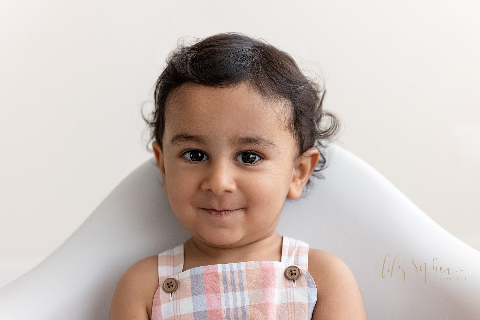  Close-up portrait of an Indian baby boy smirking proudly as he sits on his own in a white molded chair in a natural light studio near Vinings in Atlanta. 