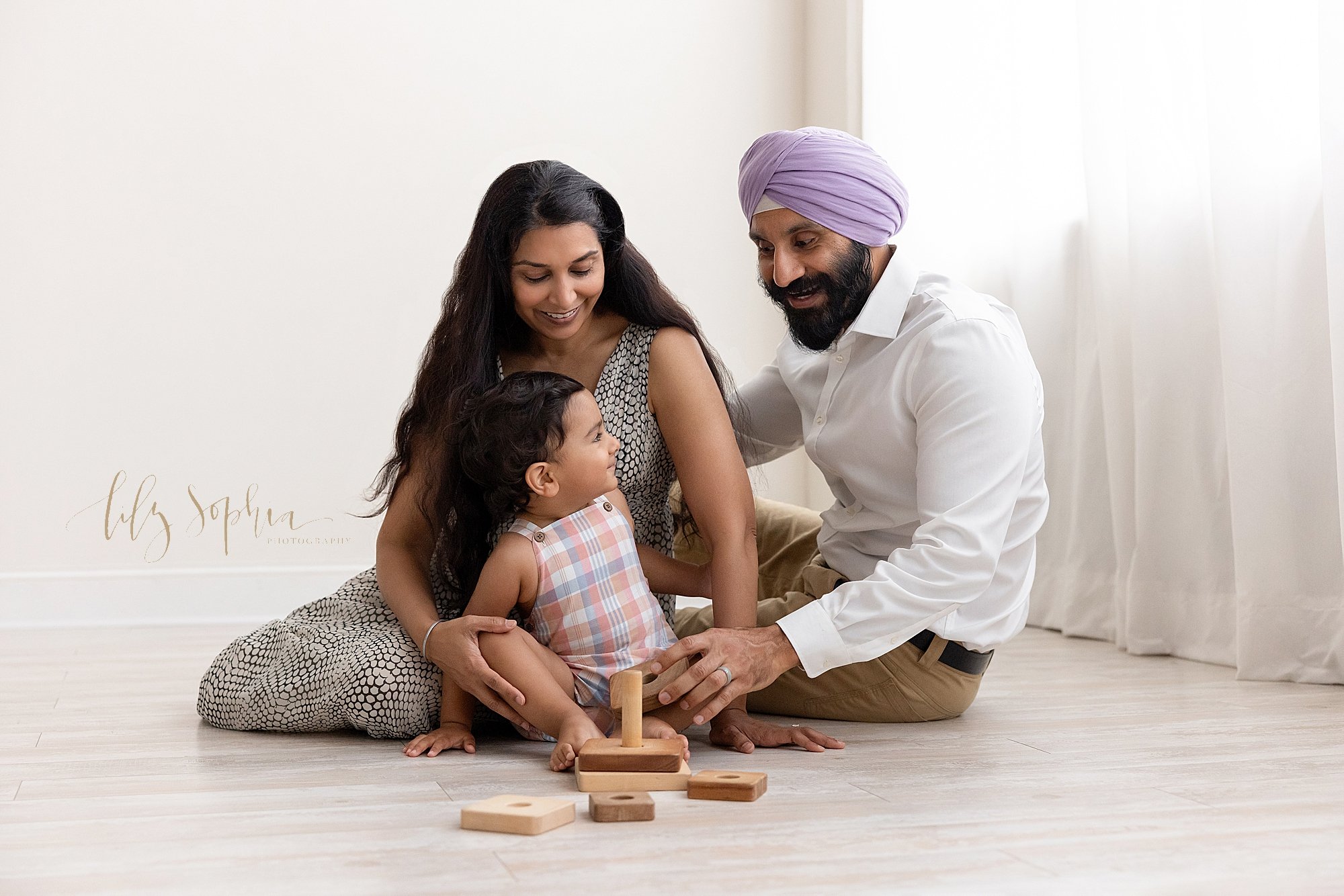  Family portrait of a mother sitting next to her husband on a floor next to a window streaming natural light with her toddler son leaning against her left leg as the family interacts while playing with a wooden stacking toy taken near Alpharetta in A