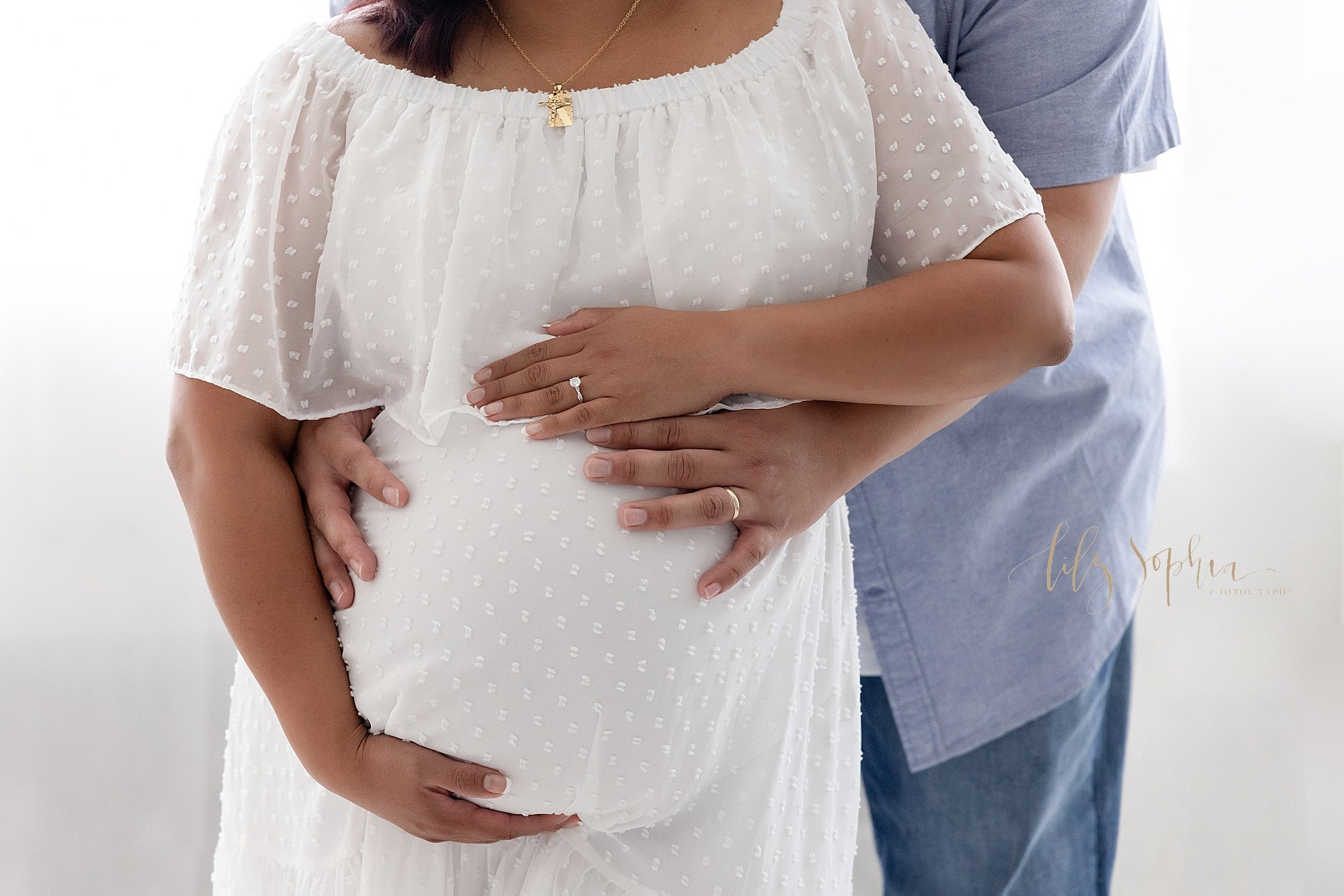  Close-up of  pregnant woman’s belly for a maternity photo session with the mother framing her belly and her husband standing behind her with his hands wrapped around her to place his hands on her belly taken near Sandy Springs in Atlanta in a natura