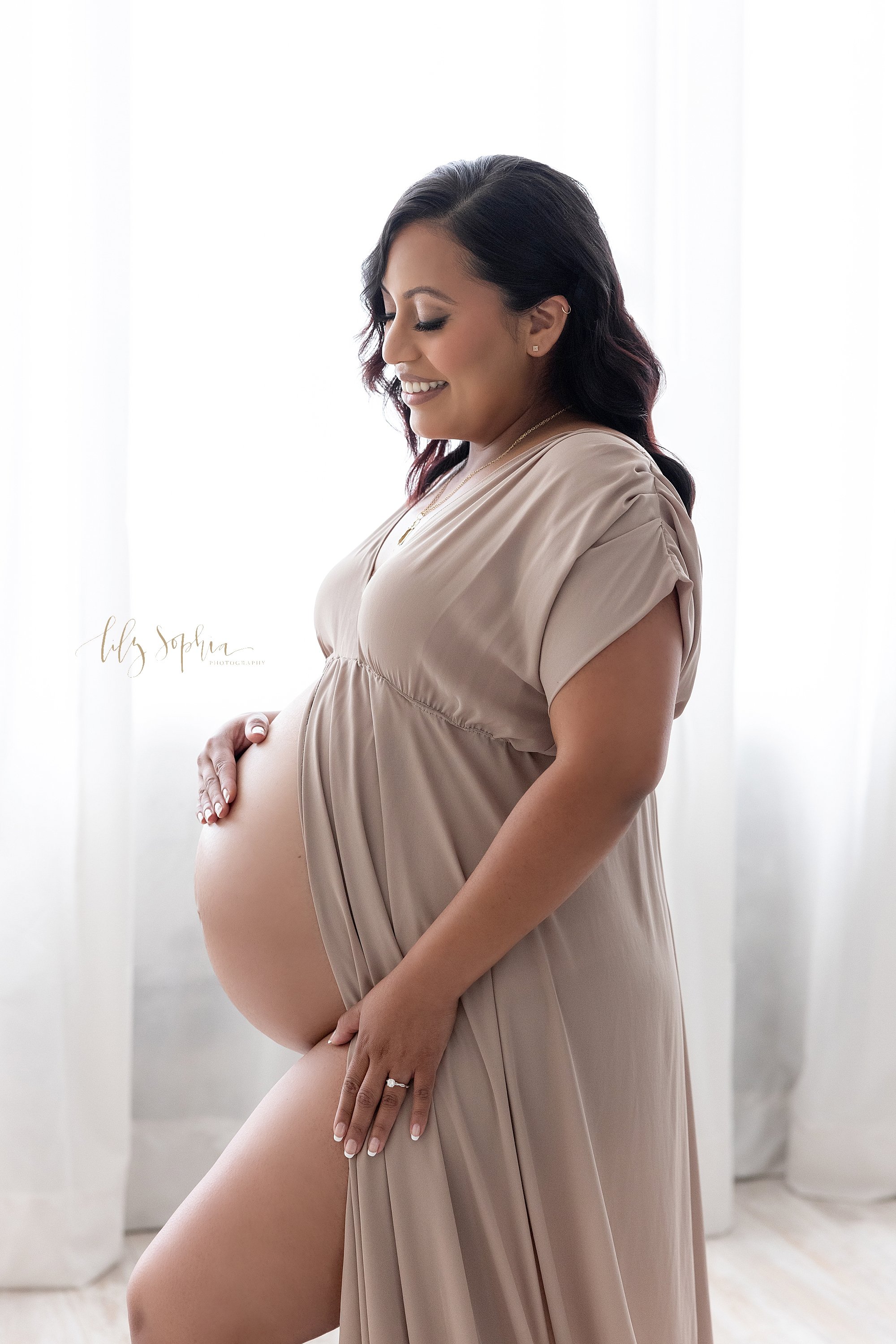  Maternity portrait of a pregnant mother standing in front of a window streaming natural light with her split front gown baring her belly as she places her right hand on her bare belly and her left hand on her gown to bare her right leg as she contem