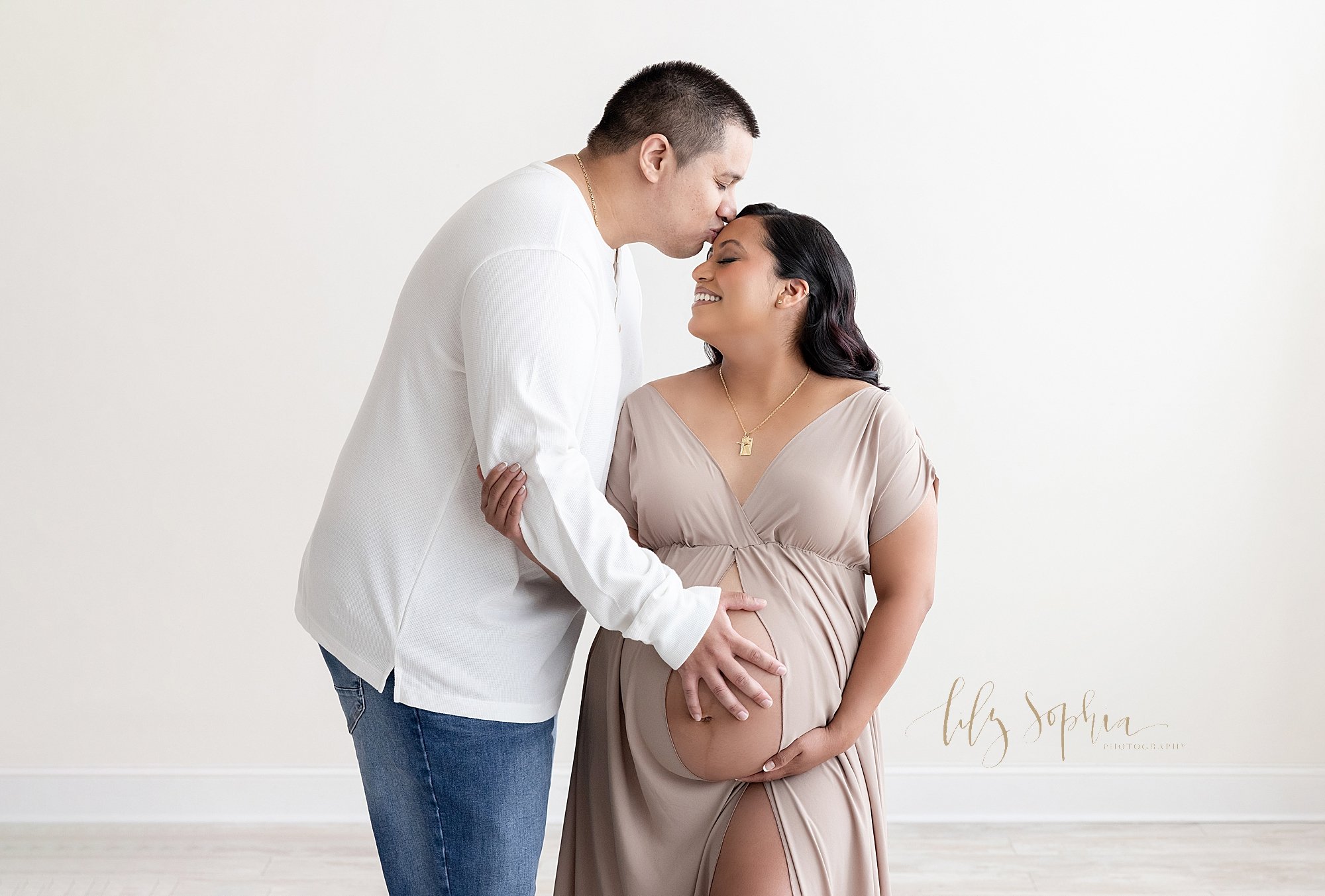  Maternity photo session with an expectant couple as the mother wears a split front full-length V-neck gown and bares her belly while her husband stands to her right side and places his right hand on their child in utero as he kisses his wife on her 