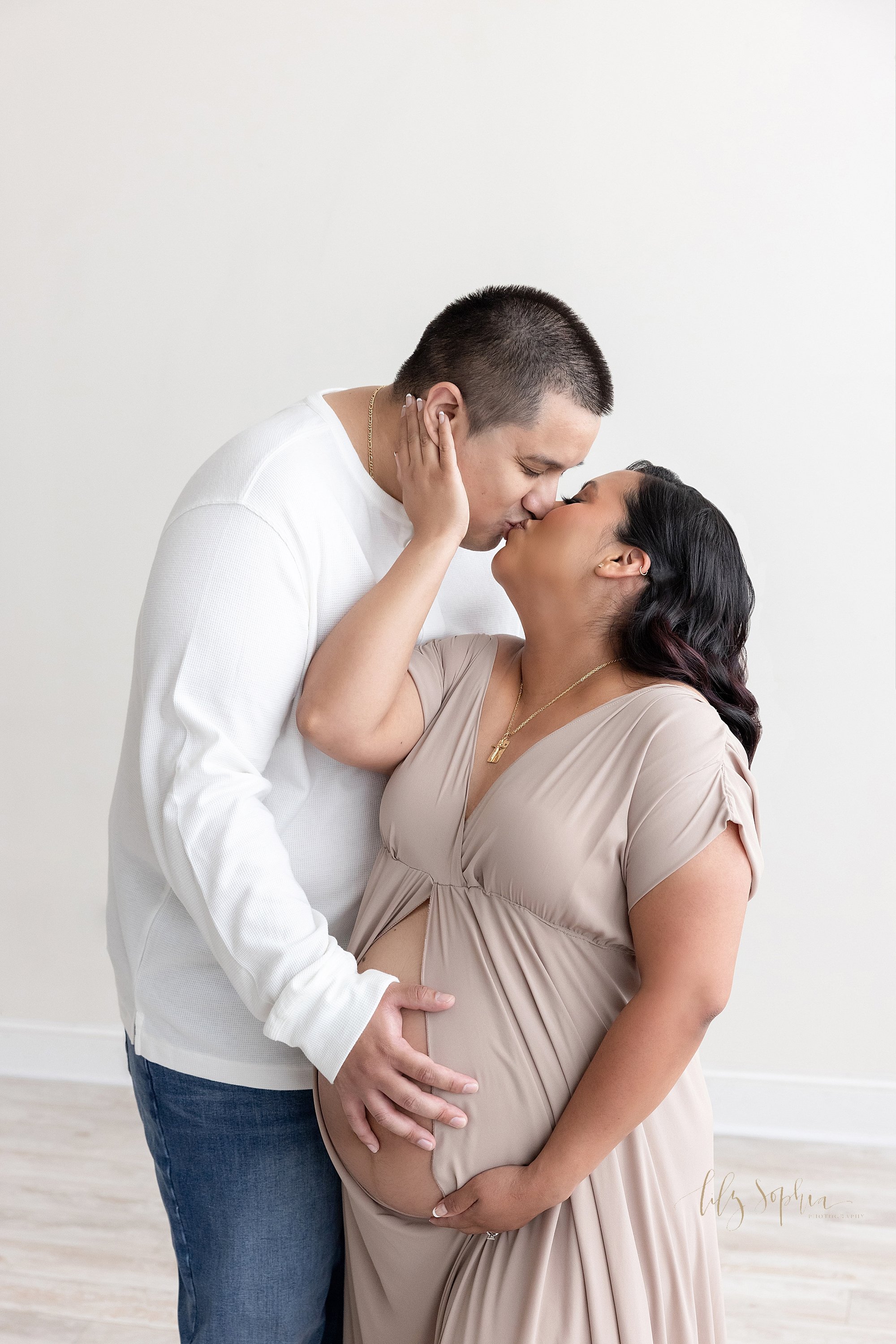  Maternity photo session with a pregnant mother standing to the left of her husband as he places his left hand on her bare belly and she pulls his face with her right hand toward her so the two of them can kiss as the couple stands in a studio near K