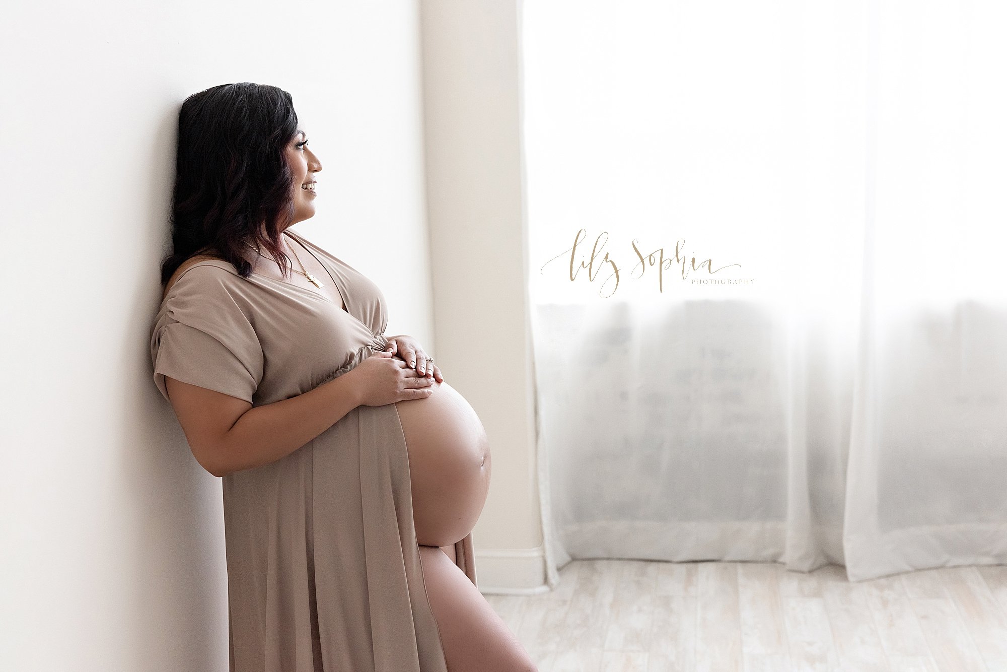  Maternity photo shoot with a pregnant mother wearing a V-neck split front jersey knit full-length gown standing with her back against a wall and her hands resting on top of her bare belly with her right leg bent as she looks out a window streaming n