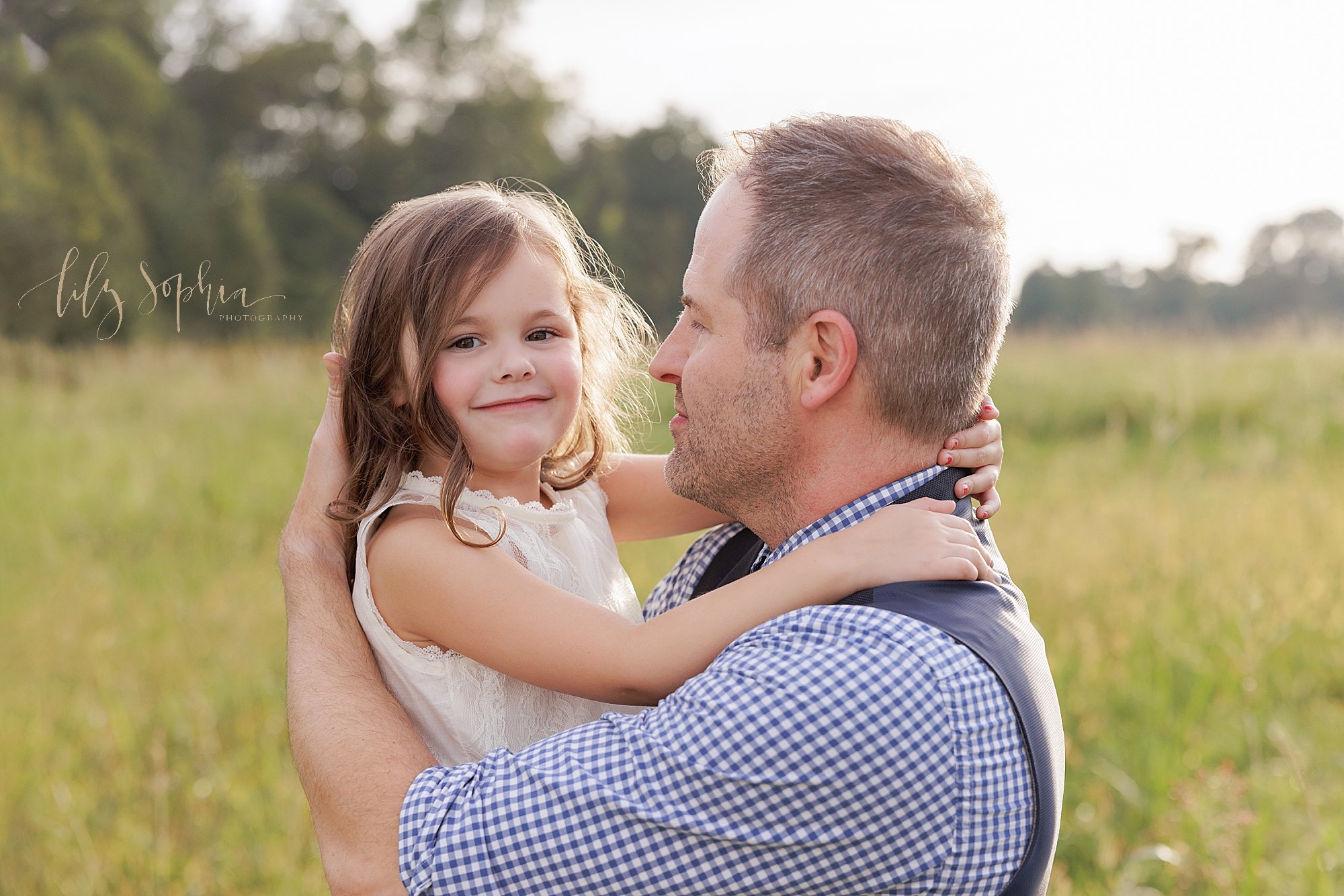  Family portrait of a young girl as she is held in her father’s arms and she holds onto his neck at sunset in a field near Atlanta. 