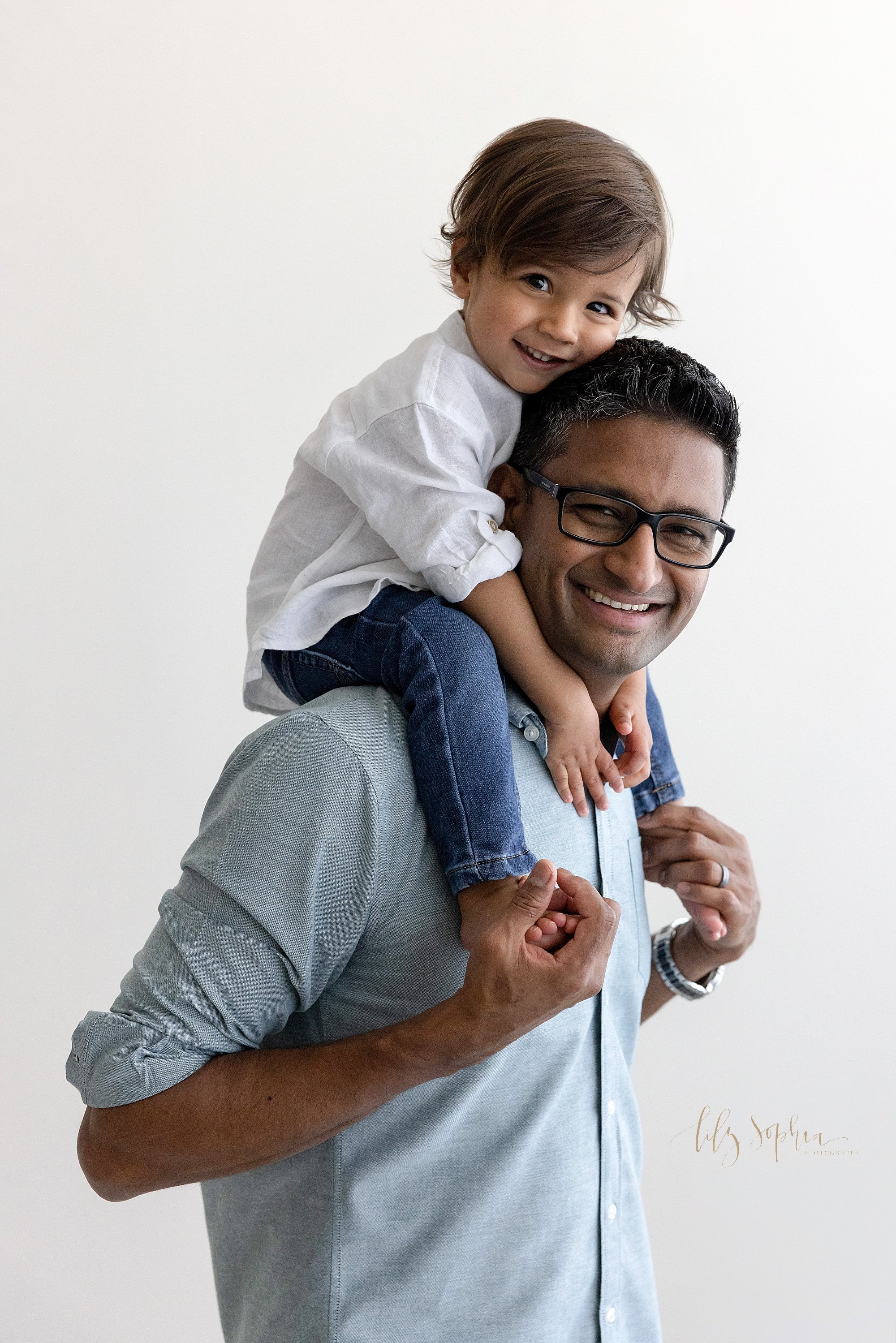  Family photo of a father giving his toddler son a piggyback ride on his shoulders as his son rests his head against his dad’s head taken next to a window that streams natural light into a photography studio in Ponce City Market in Atlanta, Georgia. 