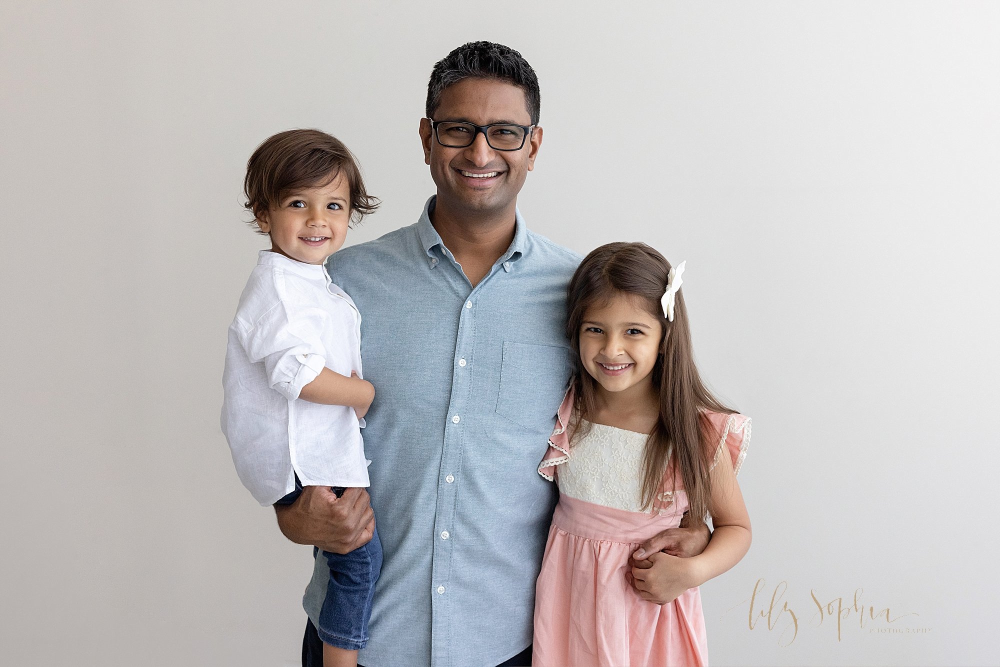  Family picture of a father holding his toddler son in his right arm as he stands in a photography studio with his young daughter on his left side as they hold hands around her waist taken in a studio near Kirkwood in Atlanta that uses natural light.