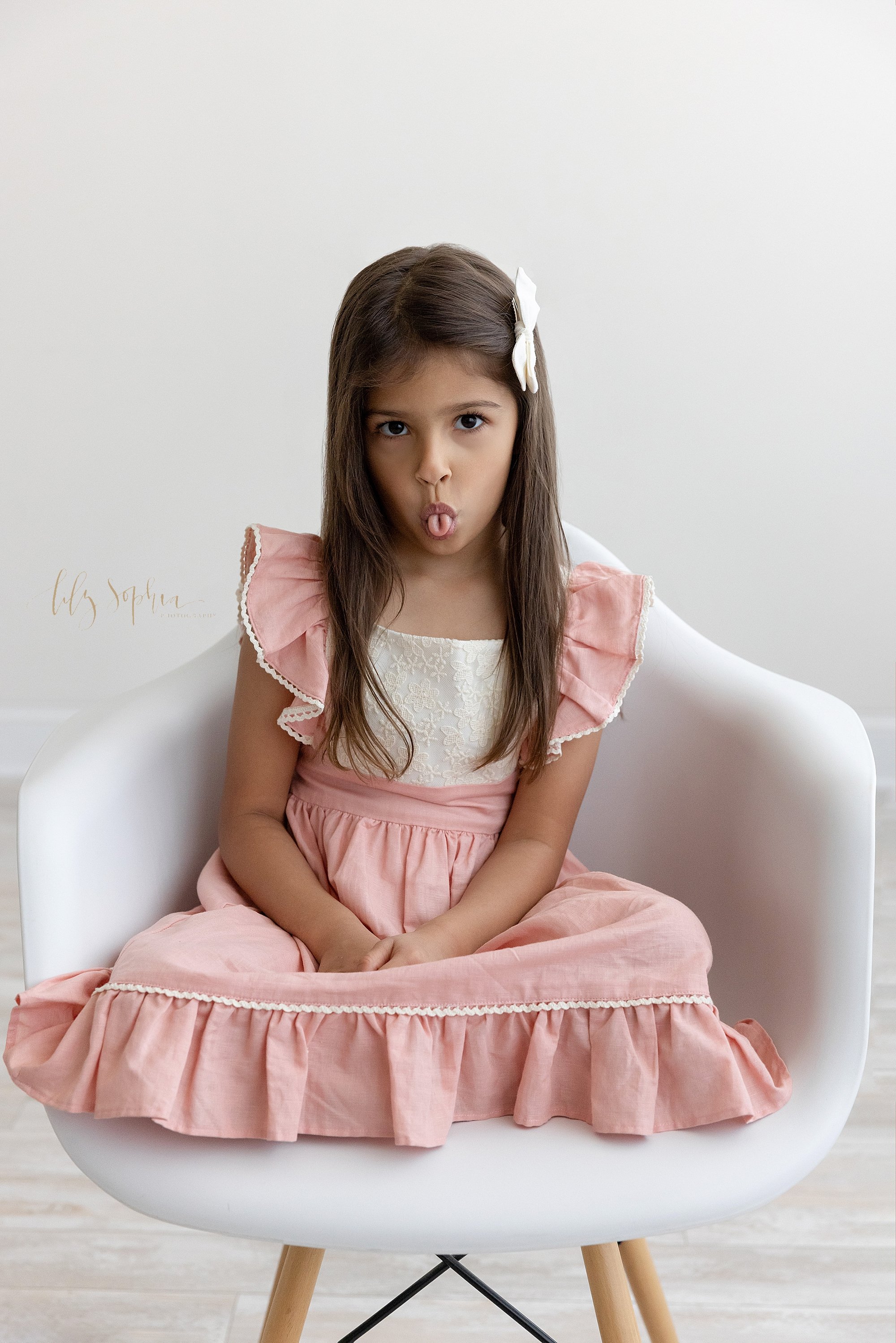  Portrait of a young girl as she sits cross legged in a white molded chair with her tongue rolled and sticking out of her mouth taken near Decatur in Atlanta in a natural light studio. 