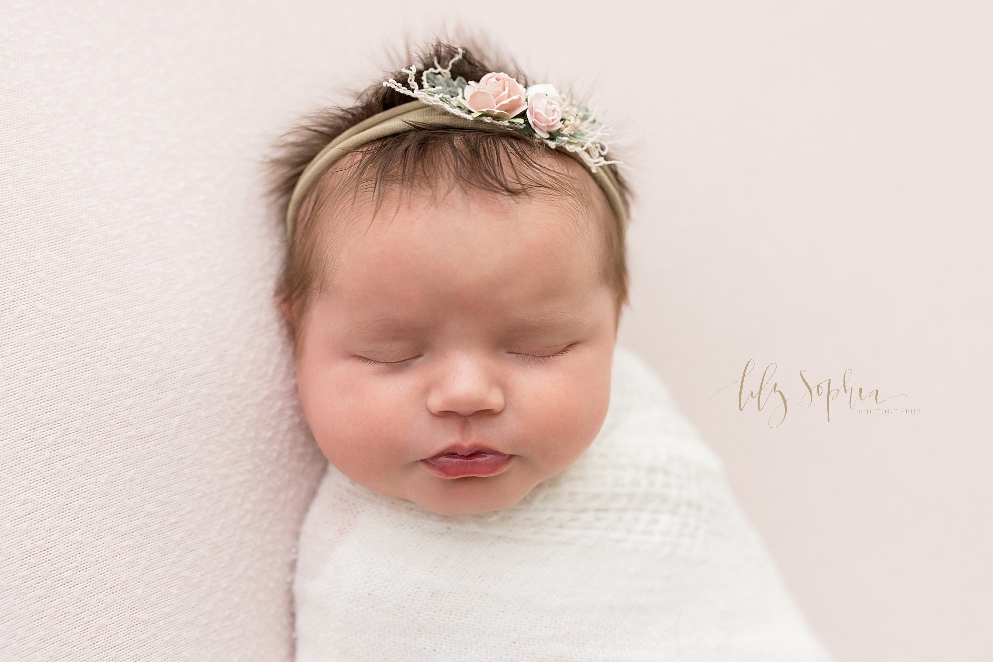 Newborn portrait of a peacefully sleeping newborn baby girl swaddled to her chin in a soft white crocheted blanket wearing a rose headband in her hair as she lies on her back featuring her wispy eyelashes, button nose and plump lips taken in natural