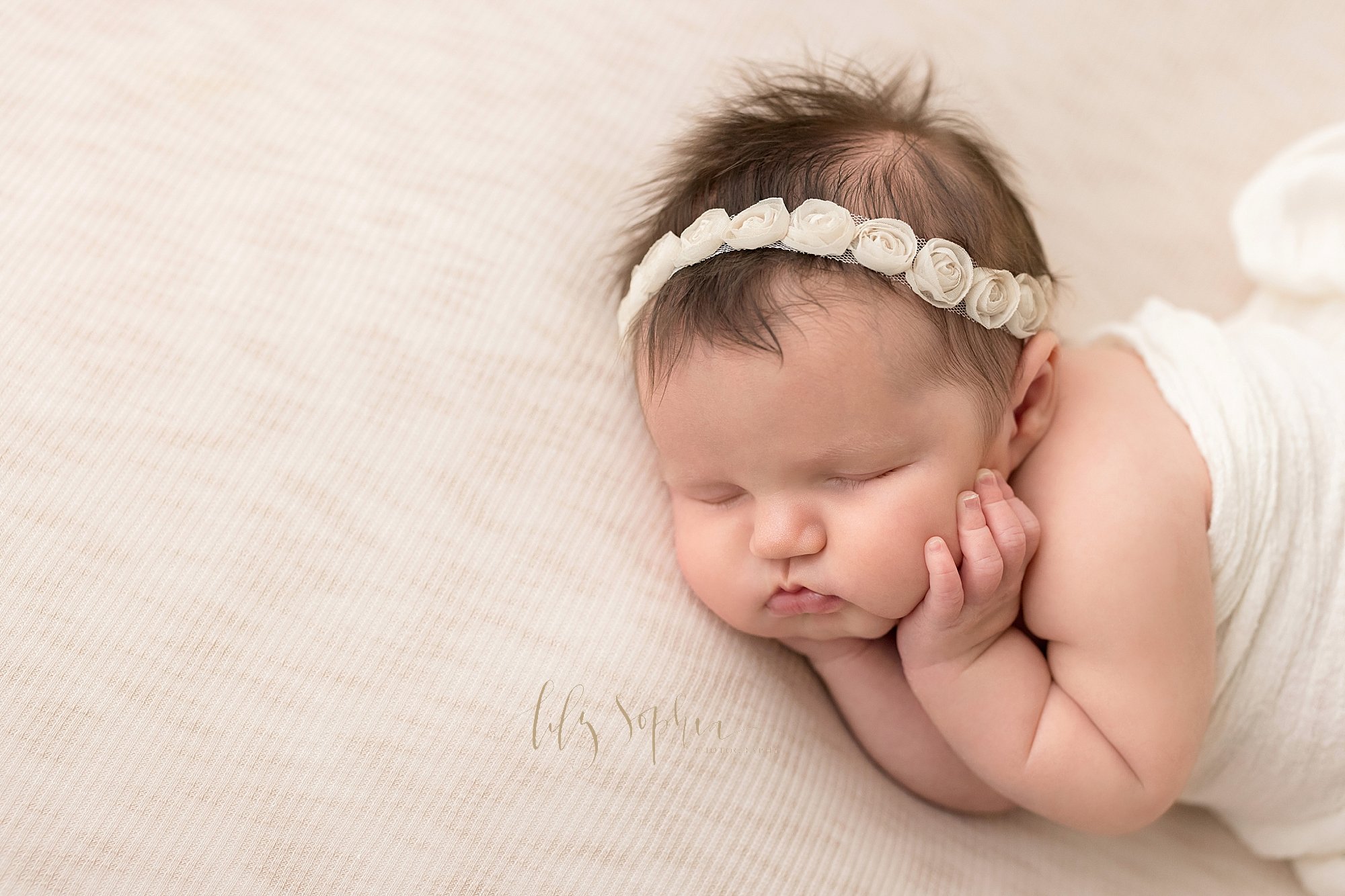  Newborn picture of a newborn baby girl wearing a tulle rose headband in her fly away hair lying on her right side with her chubby cheeks held in her hands as she sleeps in a photography studio near Smyrna in Atlanta that uses natural light. 