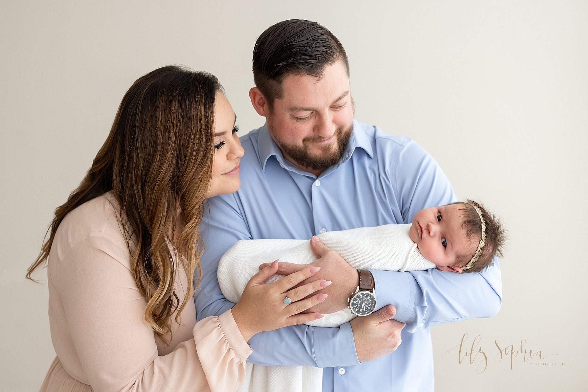  Family newborn photo of a father cradling his awake newborn baby girl in his arms as his wife stands next to him on his right side and the two of them look on while standing in a studio that uses natural light near Roswell in Atlanta, Georgia. 