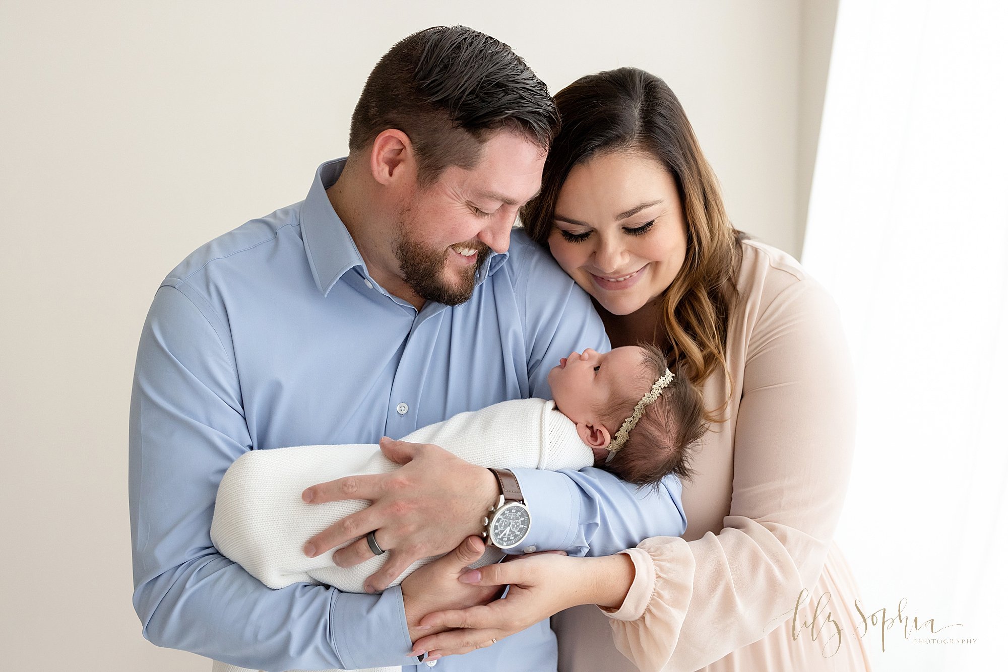  Family newborn portrait of a an awake newborn baby girl looking up at her smiling father as he cradles her in his arms and mom stands on his left side and smiles at her daughter taken next to a window streaming natural light in a photography studio 