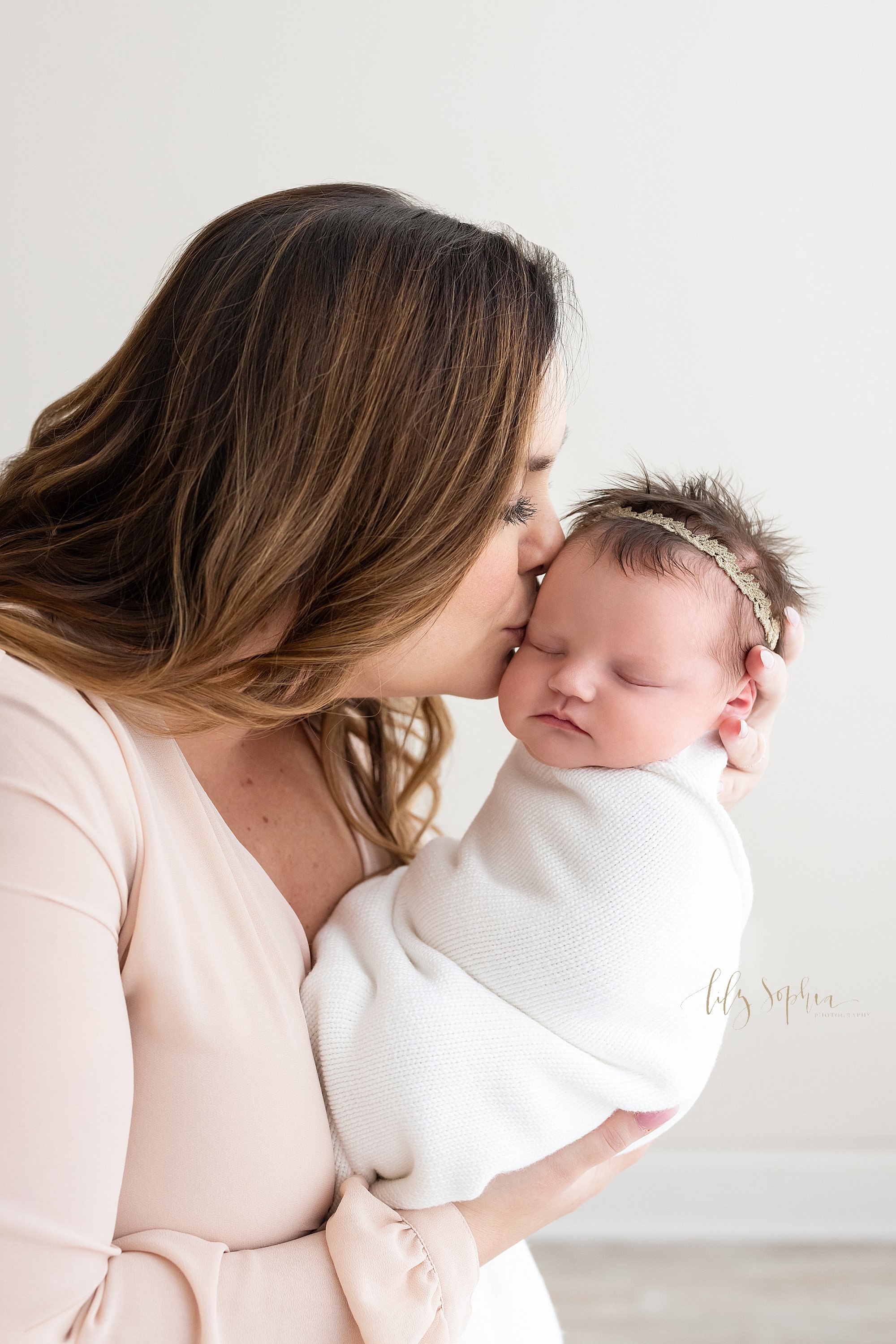  Newborn portrait of a newborn baby girl with her mom holding her head in her left hand as she kisses the side of her baby’s head taken near Old Fourth Ward in Atlanta in a studio using natural light. 