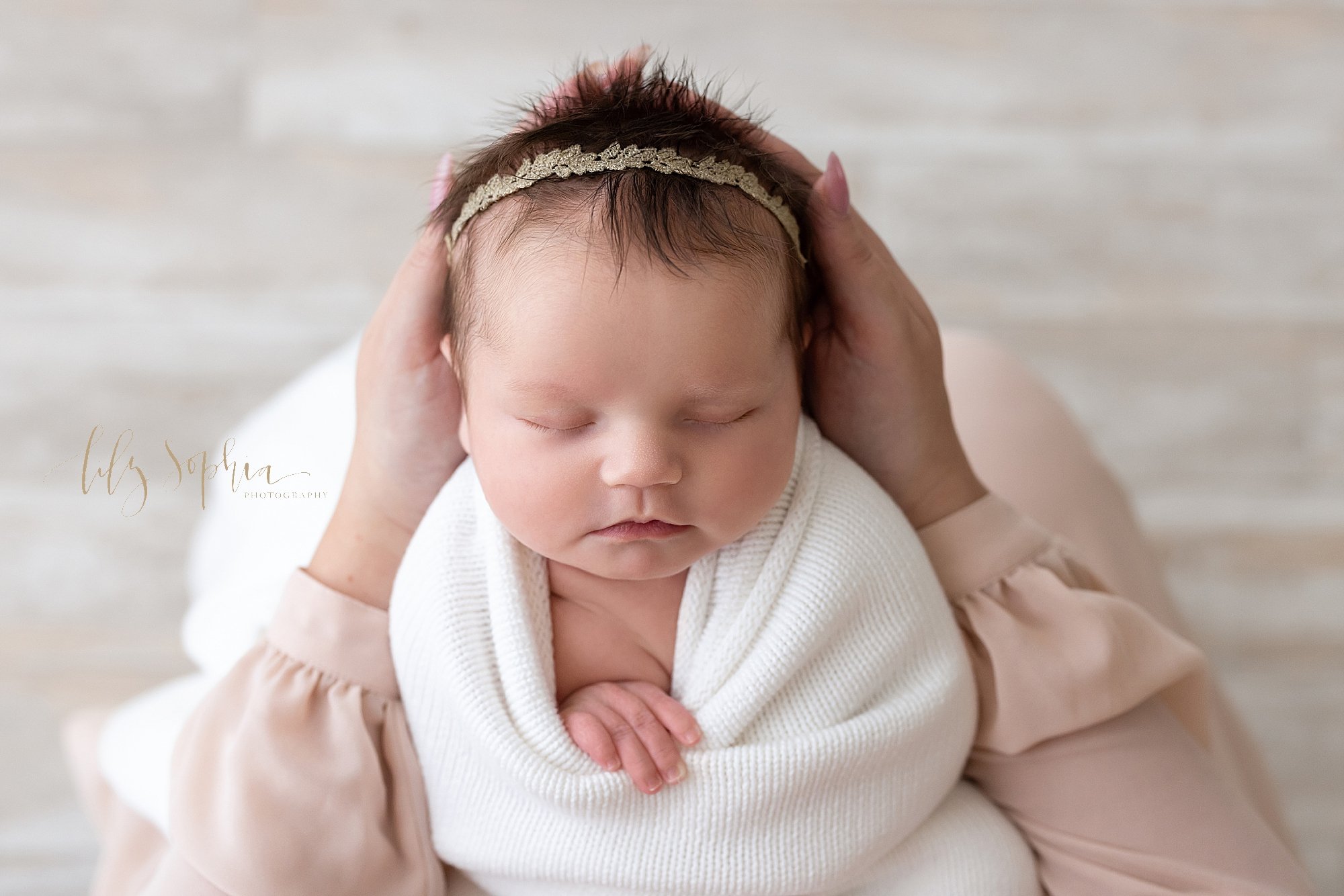 Newborn baby portrait of a newborn baby girl as she pulls her right hand out from a soft white knitted blanket swaddled around her as she sleeps peacefully in her mother’s hands taken in a natural light studio near Oakhurst in Atlanta. 