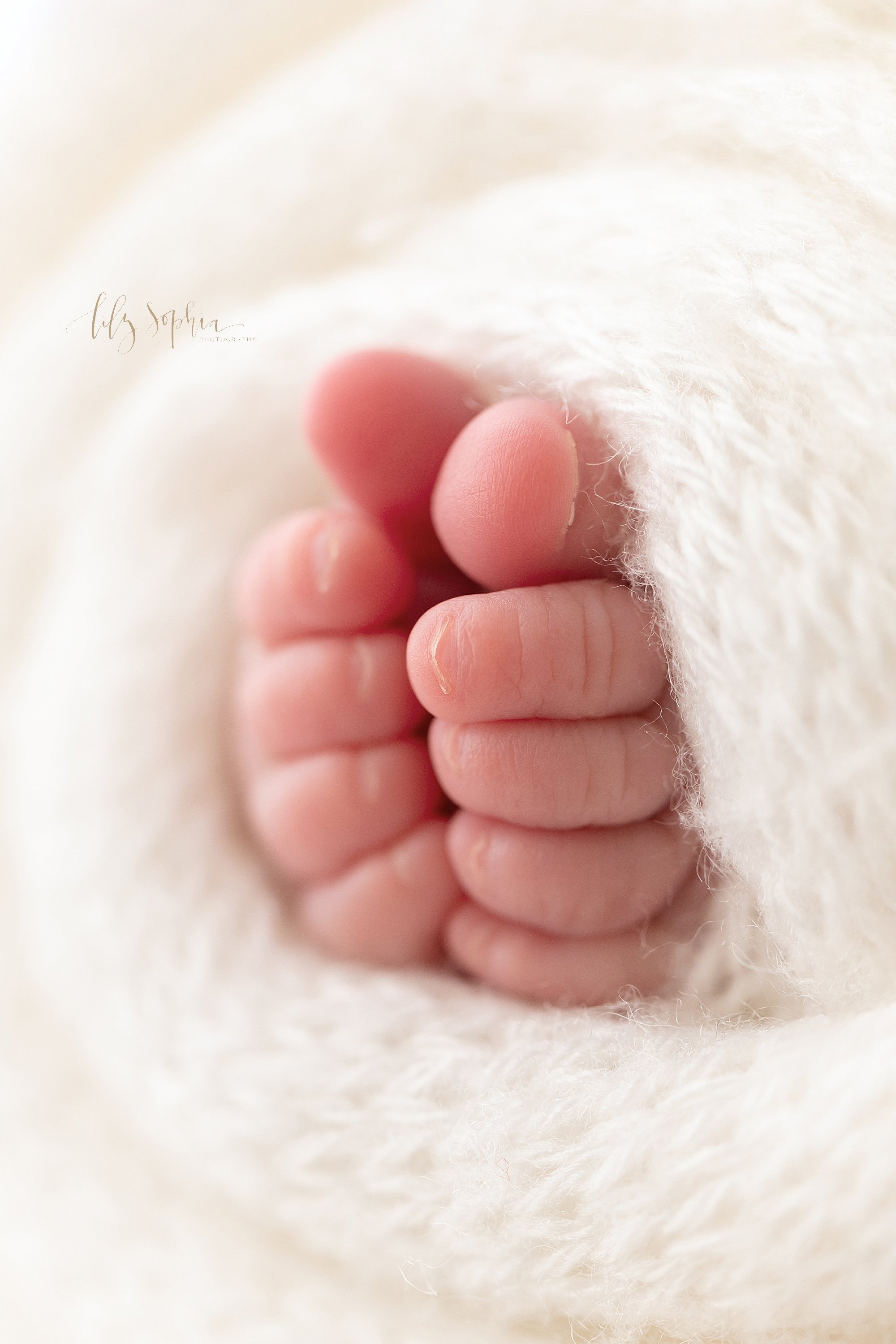  Close-up picture of a newborn baby girl’s toes peeking out from a soft white knitted blanket taken in a studio using natural light near Ansley Park in Atlanta, Georgia. 