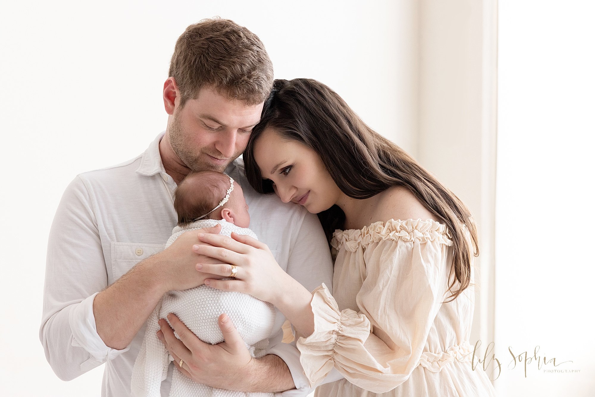  Newborn family portrait as dad holds his newborn baby girl against her chest and mom places her head on her husband’s left shoulder and the two of them place their hands on their daughter’s back as the stand next to a window streaming natural light 