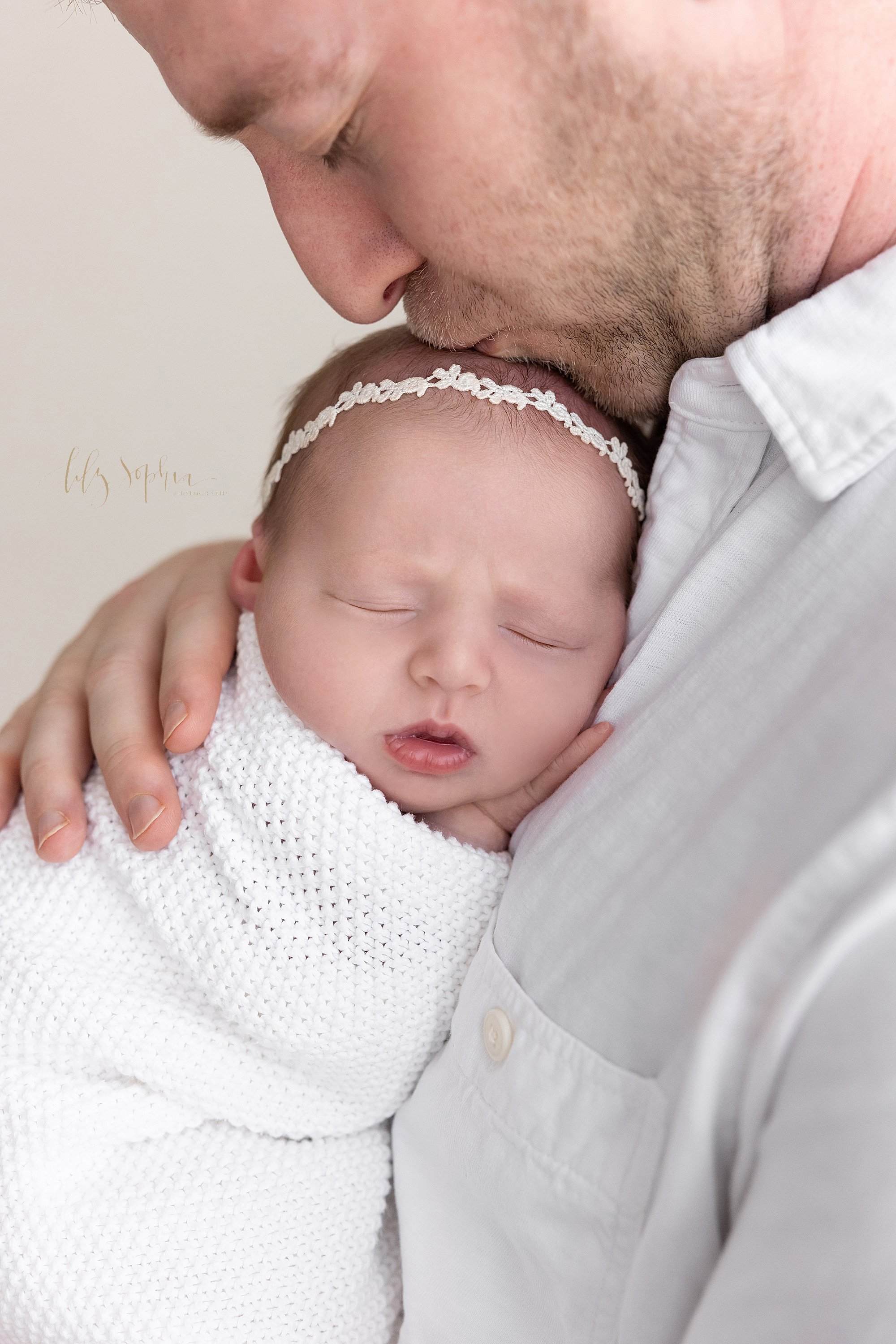  Close-up newborn photo of a newborn baby girl wrapped in a soft crocheted blanket and wearing a delicate headband in her wispy hair as her father holds her against his chest and kisses the top of her head taken in a natural light studio near Kirkwoo