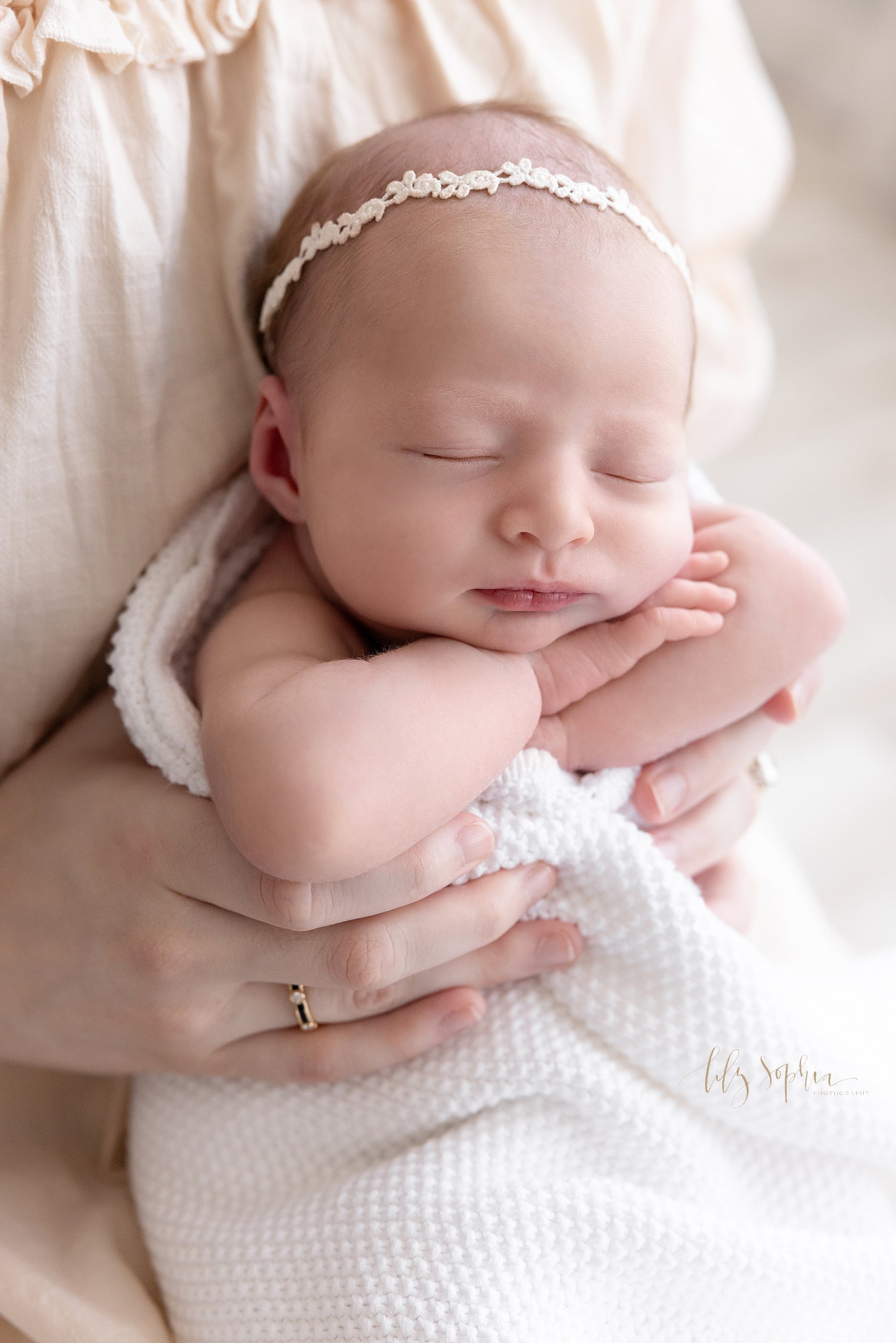  A peacefully sleeping newborn baby girl is held under her arms as she sits on her mother’s lap and places her hands on top of her arms that are folded under her chin taken using natural light near Sandy Springs in Atlanta in a photography studo. 