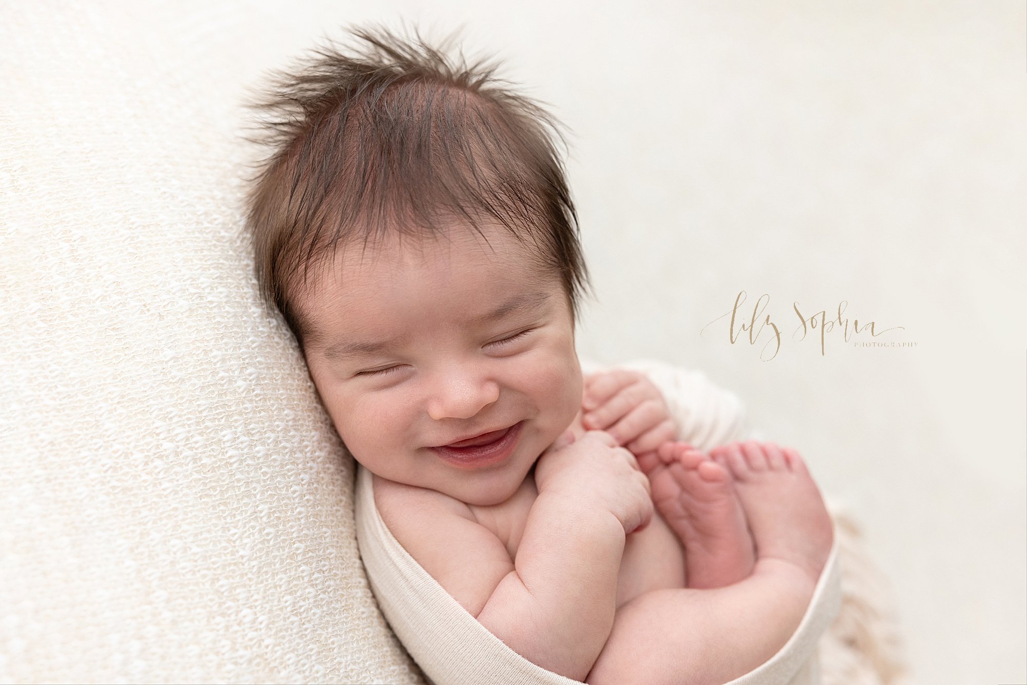  Newborn portrait of a sleeping newborn baby boy cradled in a stretchy swaddle as he smiles while lying on his back and looking over his right shoulder taken near Morningside in Atlanta in a natural light studio. 