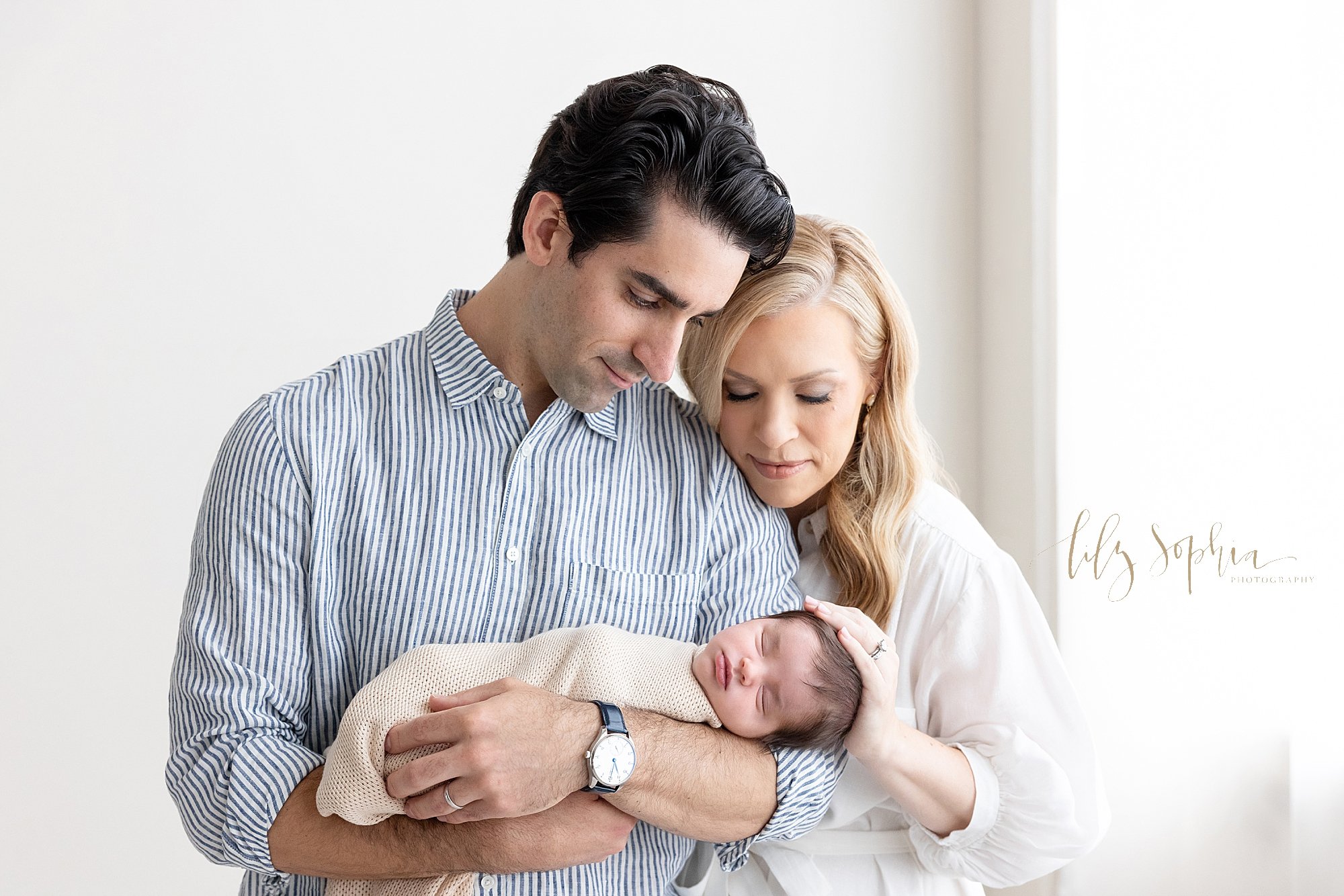  Newborn family shoot of a father cradling his peacefully sleeping newborn baby boy in his arms as his wife stands behind him on his left side with her chin against his shoulder and places her left hand on their son’s head as the couple stand next to