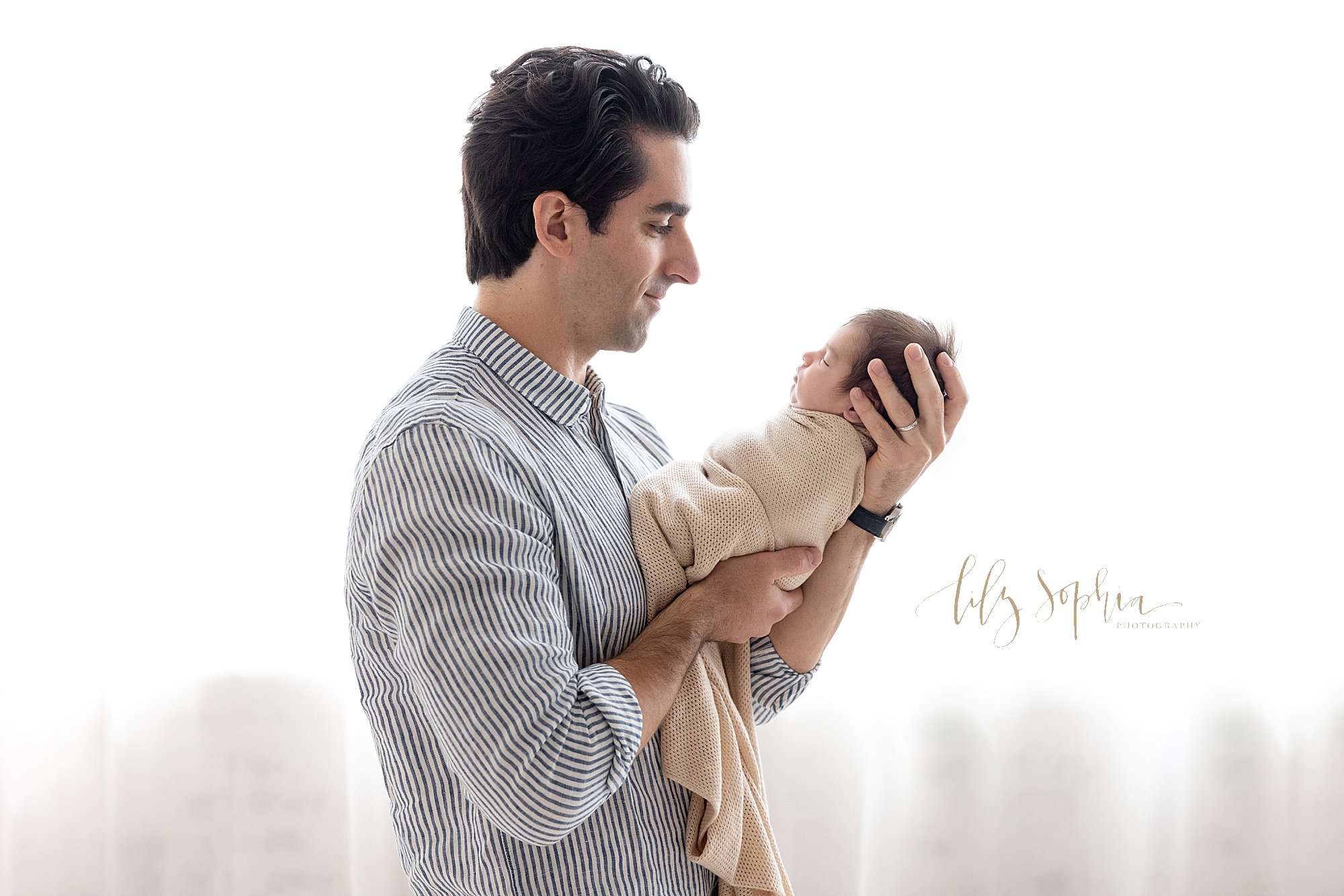  Newborn portrait of a newborn baby boy being held in front of his father as he admires him in front of a window streaming natural light in to a photography studio near Cumming in Atlanta. 