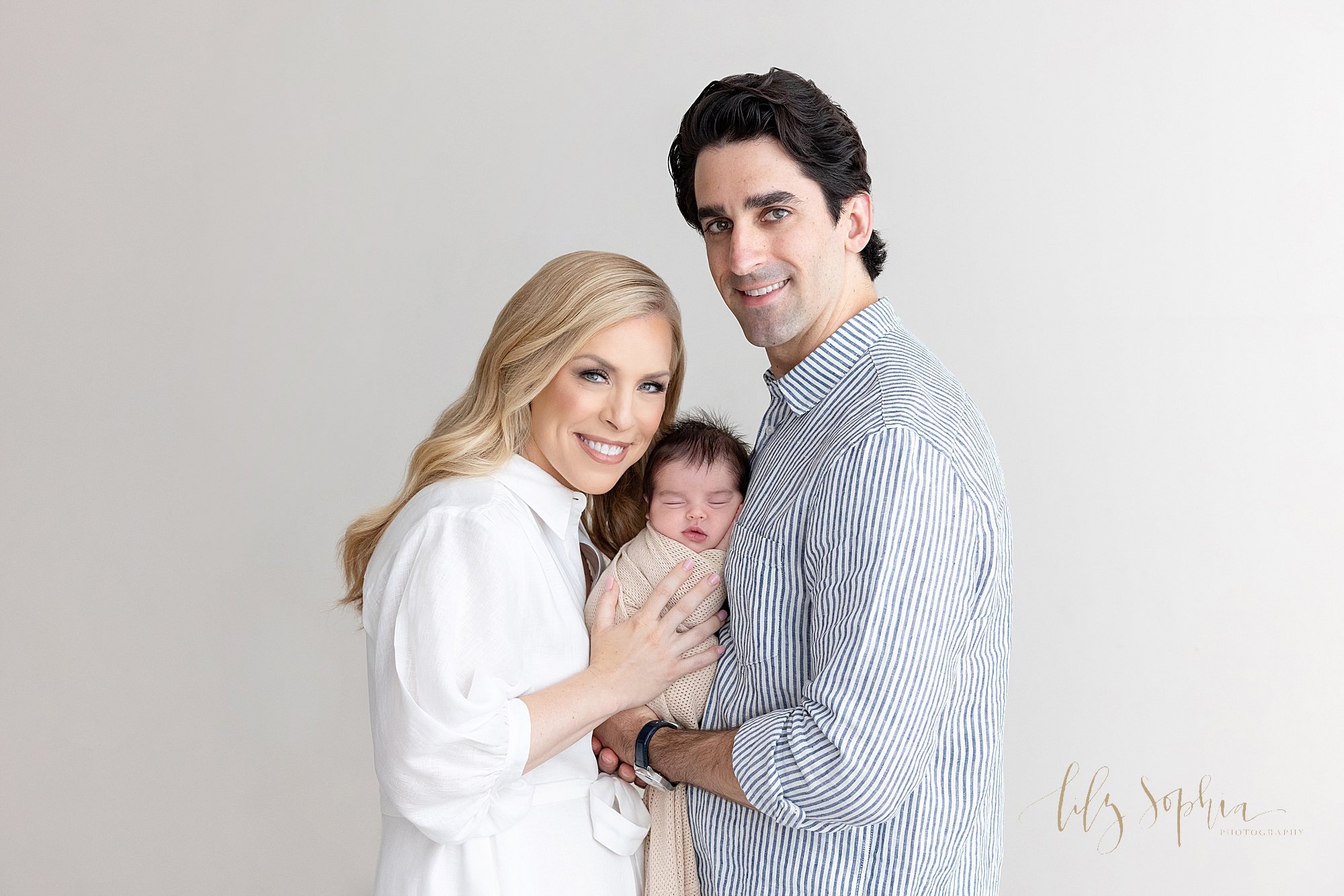 Newborn photo session in a natural light studio with a father holding his peacefully sleeping newborn against his chest while his wife faces him and places her hands against their newborn baby boy’s body and the two of them turn their heads over the