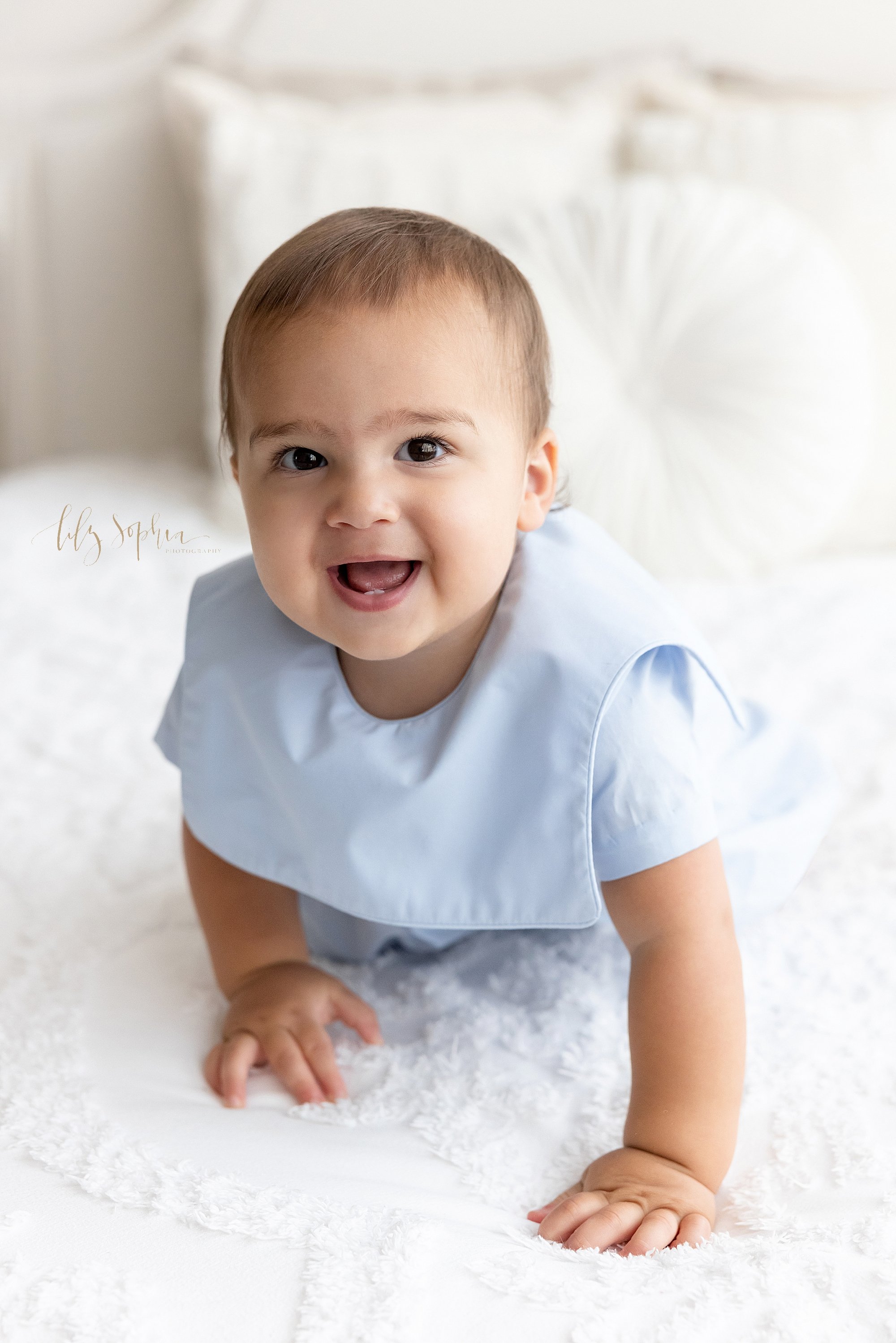  First birthday photo of a smiling one year old baby boy as he crawls across a bed in a natural light studio near Buckhead in Atlanta, Georgia. 