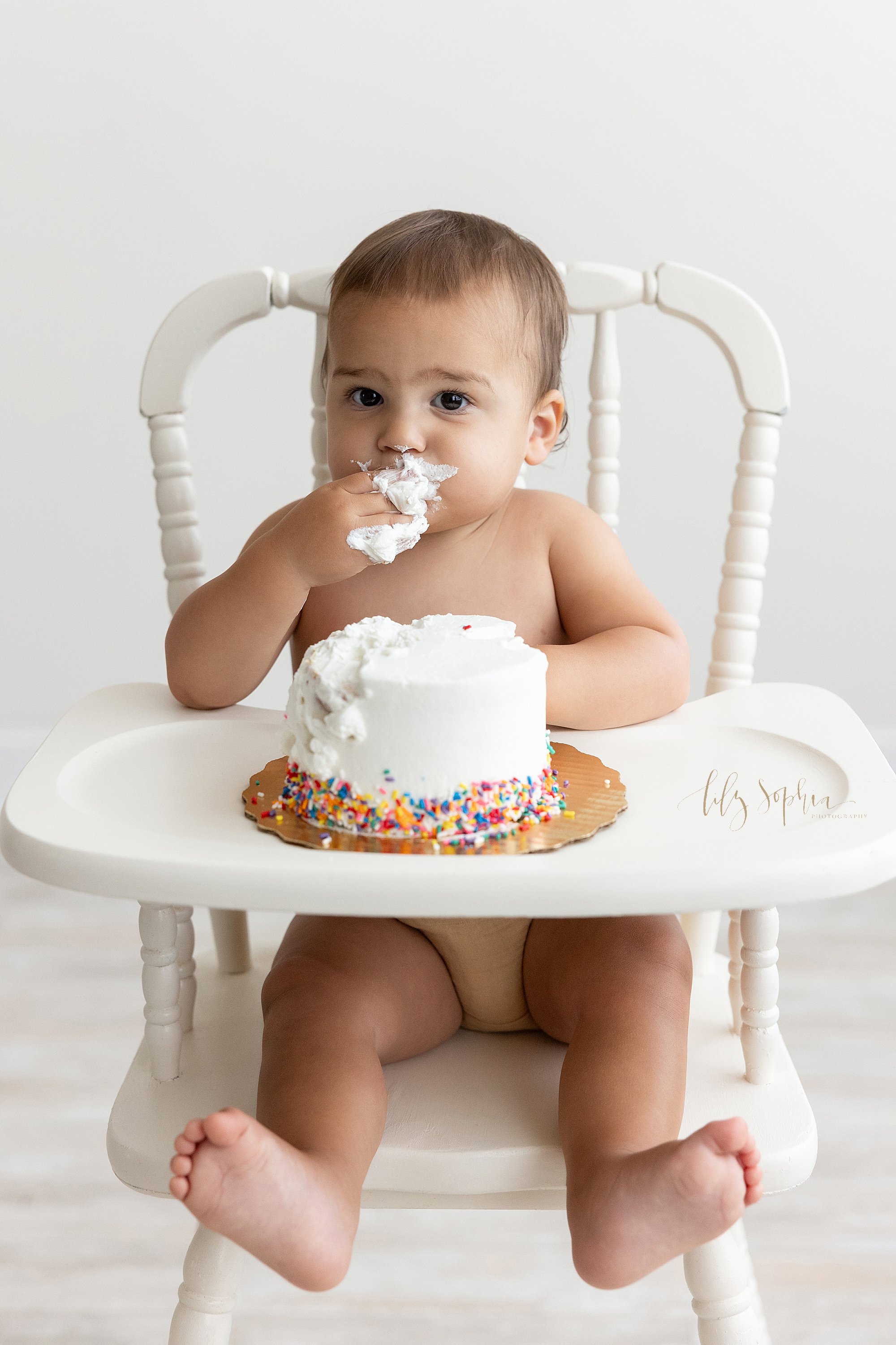  First birthday smash cake photo session of a one year old baby boy as he sits in an antique high chair shoving icing into his mouth taken next to a window streaming natural light into a photography studio near Midtown in Atlanta. 