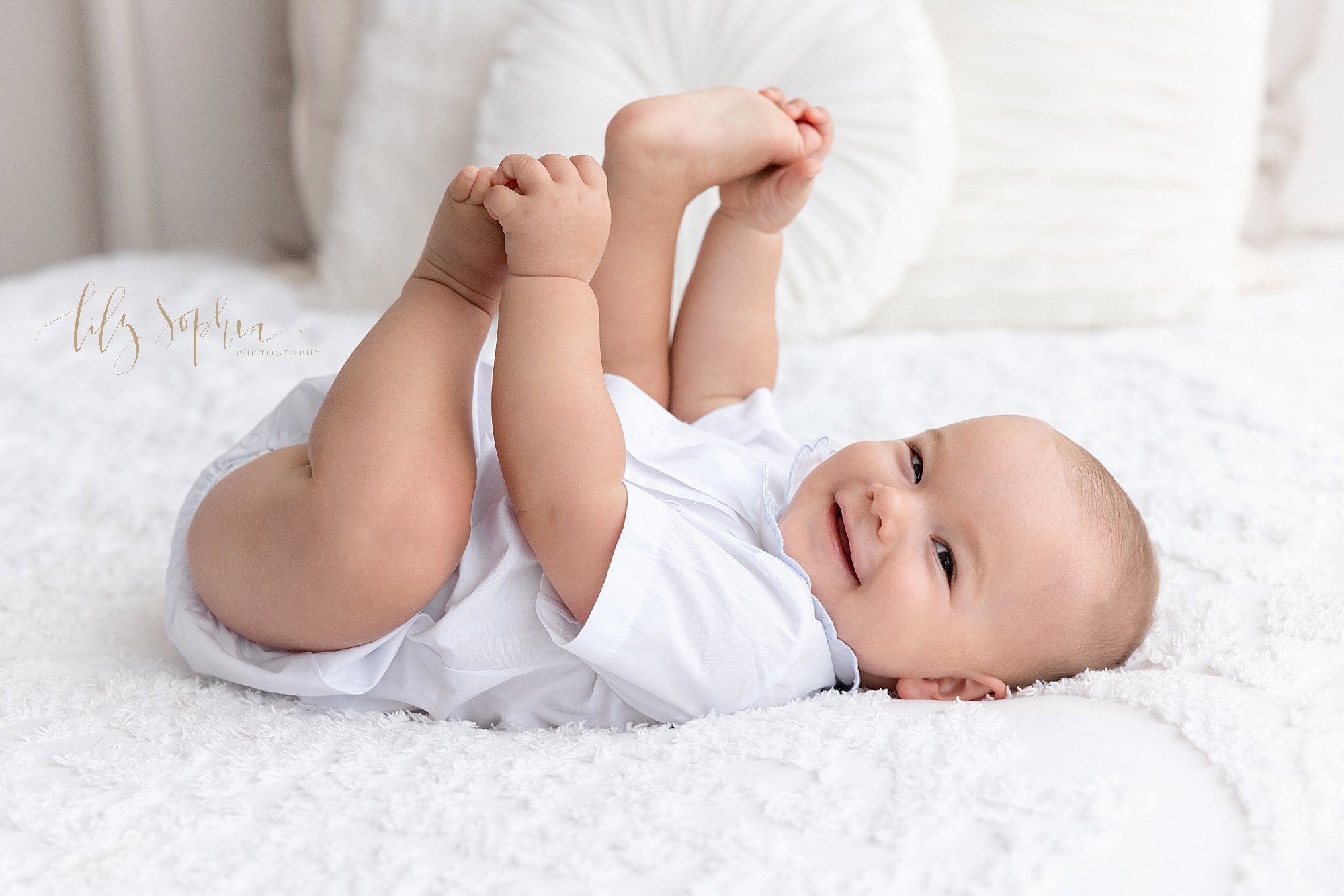  First birthday photo session of a baby boy as he lies on his back on a bed and plays with his feet taken near Alpharetta in Atlanta in a studio that uses natural light. 