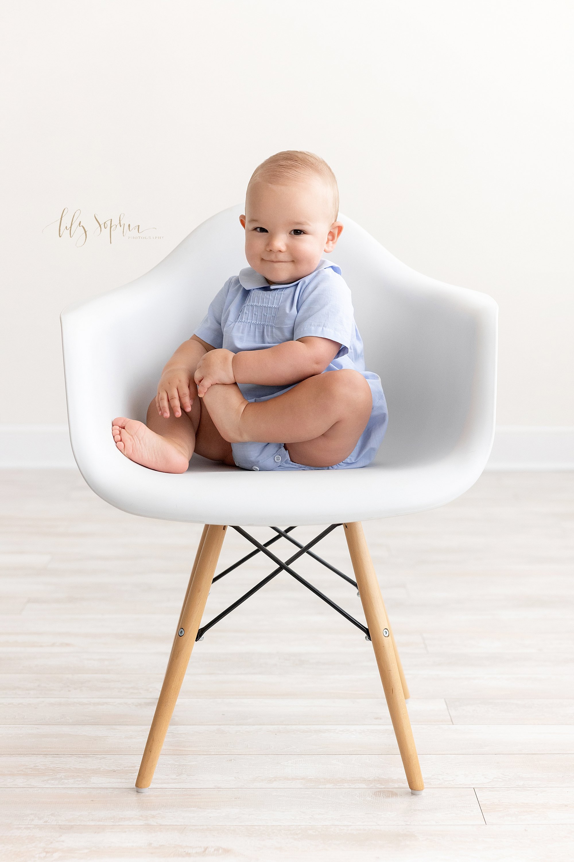 First birthday photo of a shy one year old boy as he sits in a white molded chair and grabs his left foot with his left hand taken near Brookhaven in Atlanta in a natural light studio. 