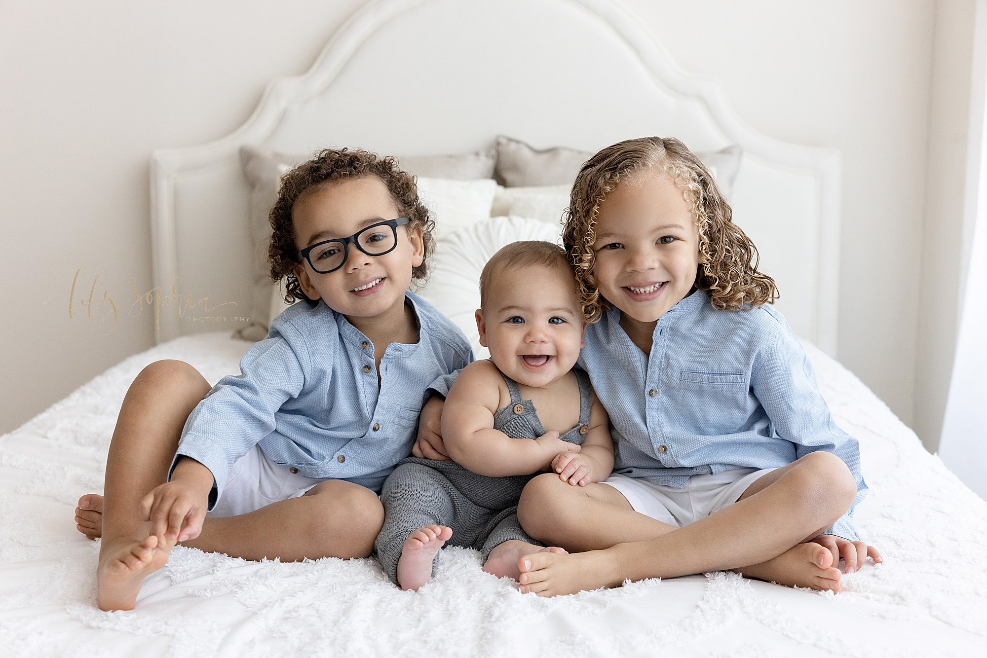  Family photo of siblings sitting on top of a bed in a natural light studio, a smiling baby boy sitting between his two happy brothers taken near Alpharetta in Atlanta, Georgia. 