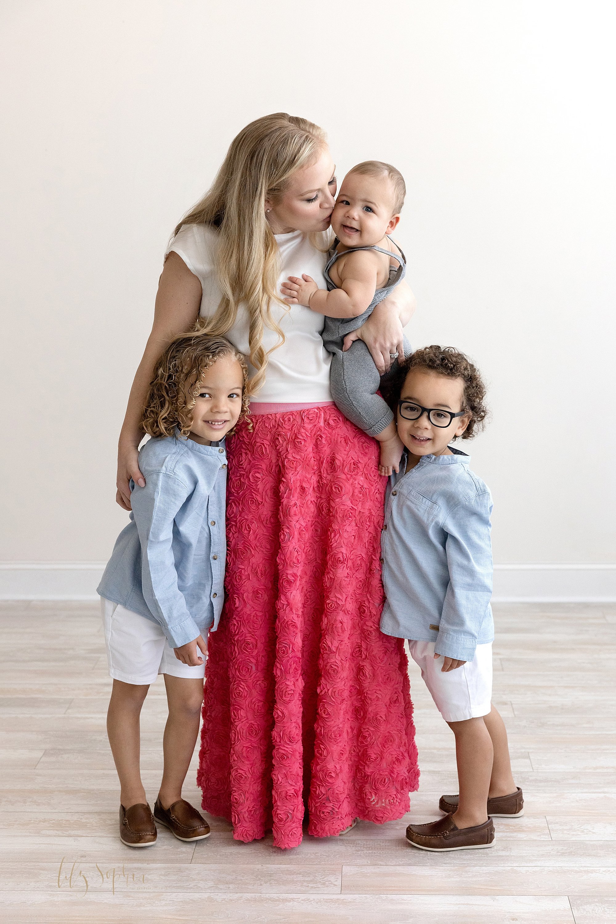  Family photograph of a mother and her sons with mom holding her baby boy with her left arm while kissing him on the cheek as she holds her oldest son with her right arm and her other son stands on his mom’s left side taken in a studio using natural 